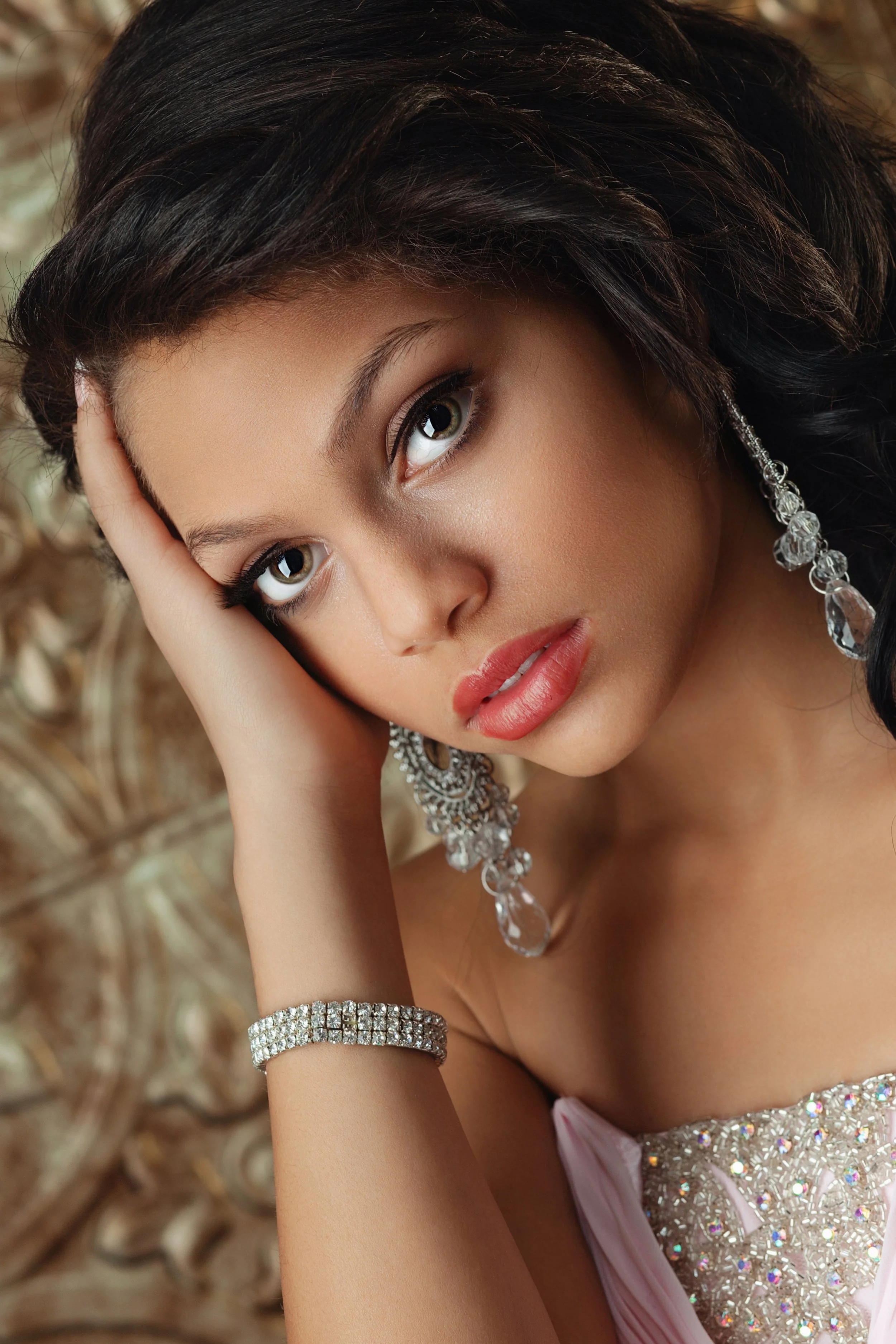 Close-up of a woman with dark hair, wearing large, sparkling earrings and a bracelet, resting her head on her hand, dressed in a pink outfit with decorative embellishments.