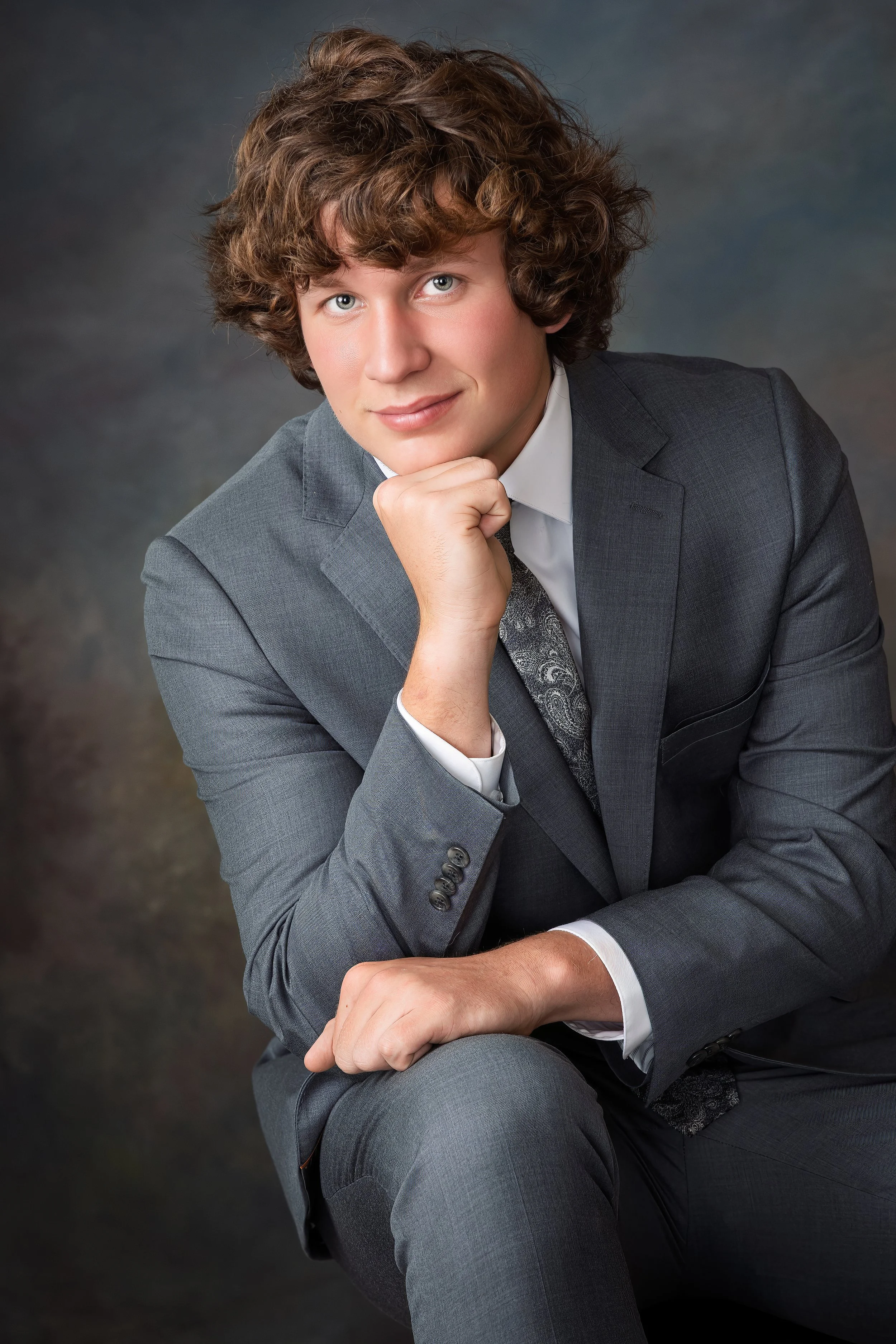 A Senior Portrait of a young man with curly brown hair wearing a gray suit, white dress shirt, and patterned tie, sitting with his chin resting on his hand and smiling at the camera against a dark, textured background.