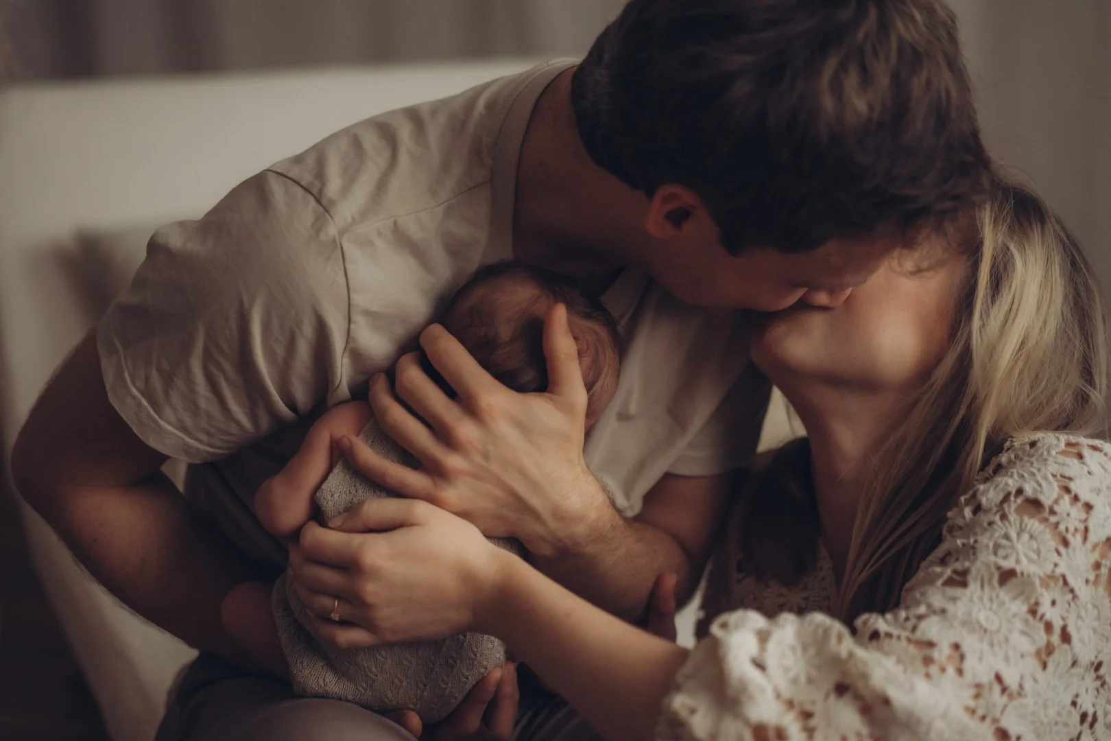 Frischgebackene Eltern küssen sich, während Papa das neugeborene Baby auf dem Arm hält.