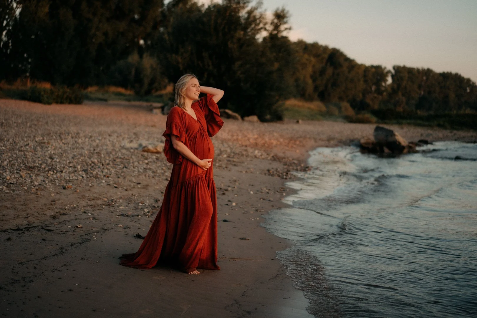 Schwangere steht am Strand im Sonnenaufgang und hält ihren Bauch.