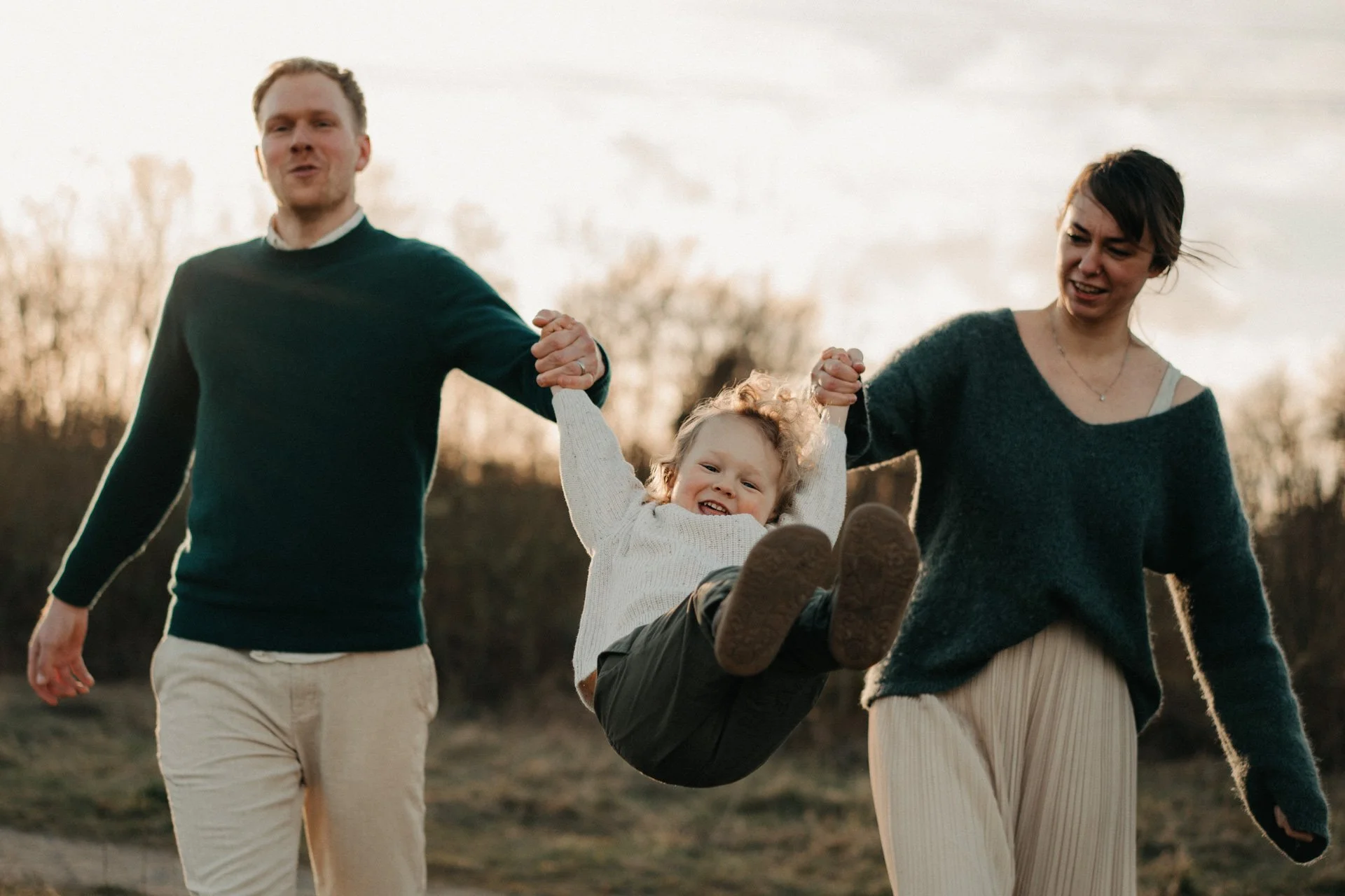 Kind schwingt an den Armen seiner Eltern auf einer Wiese.