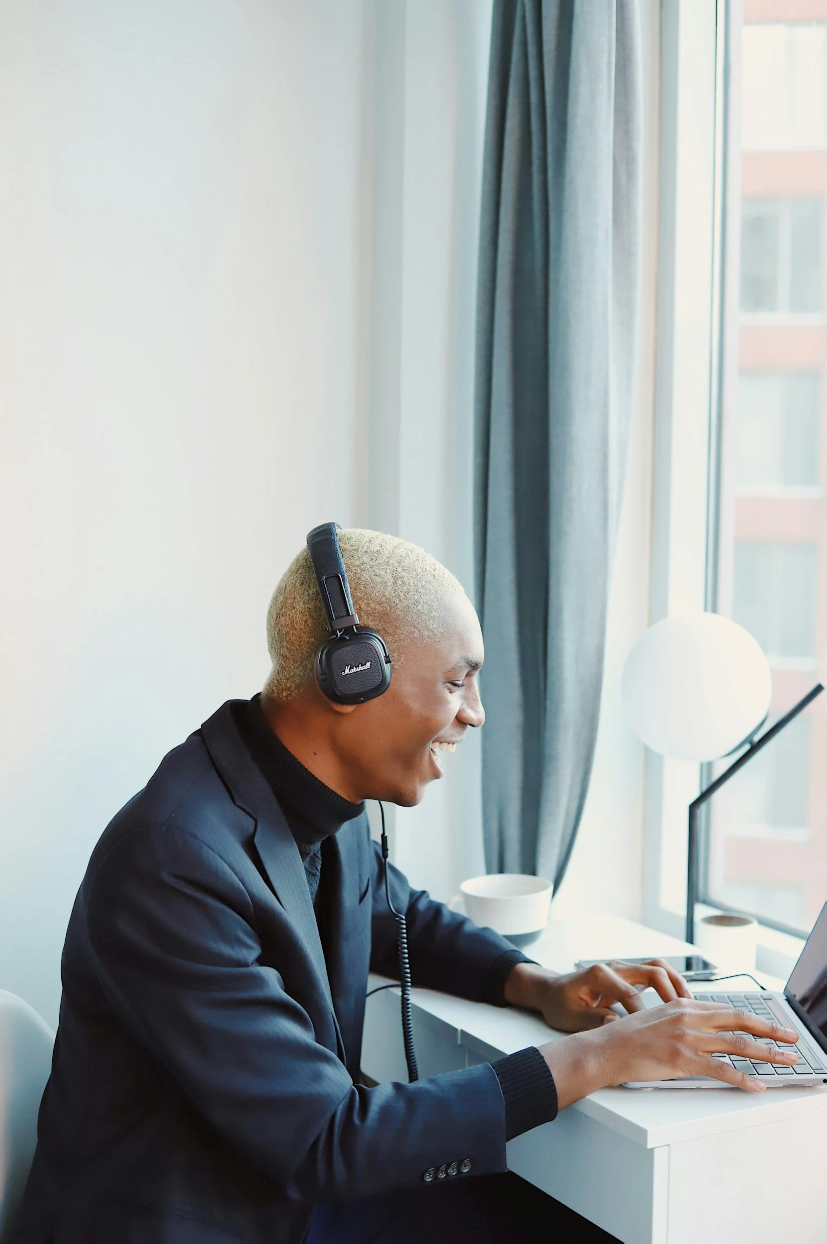 A young man with blond hair smiling and wearing a headset, sitting at a desk with a laptop near a window with gray curtains.