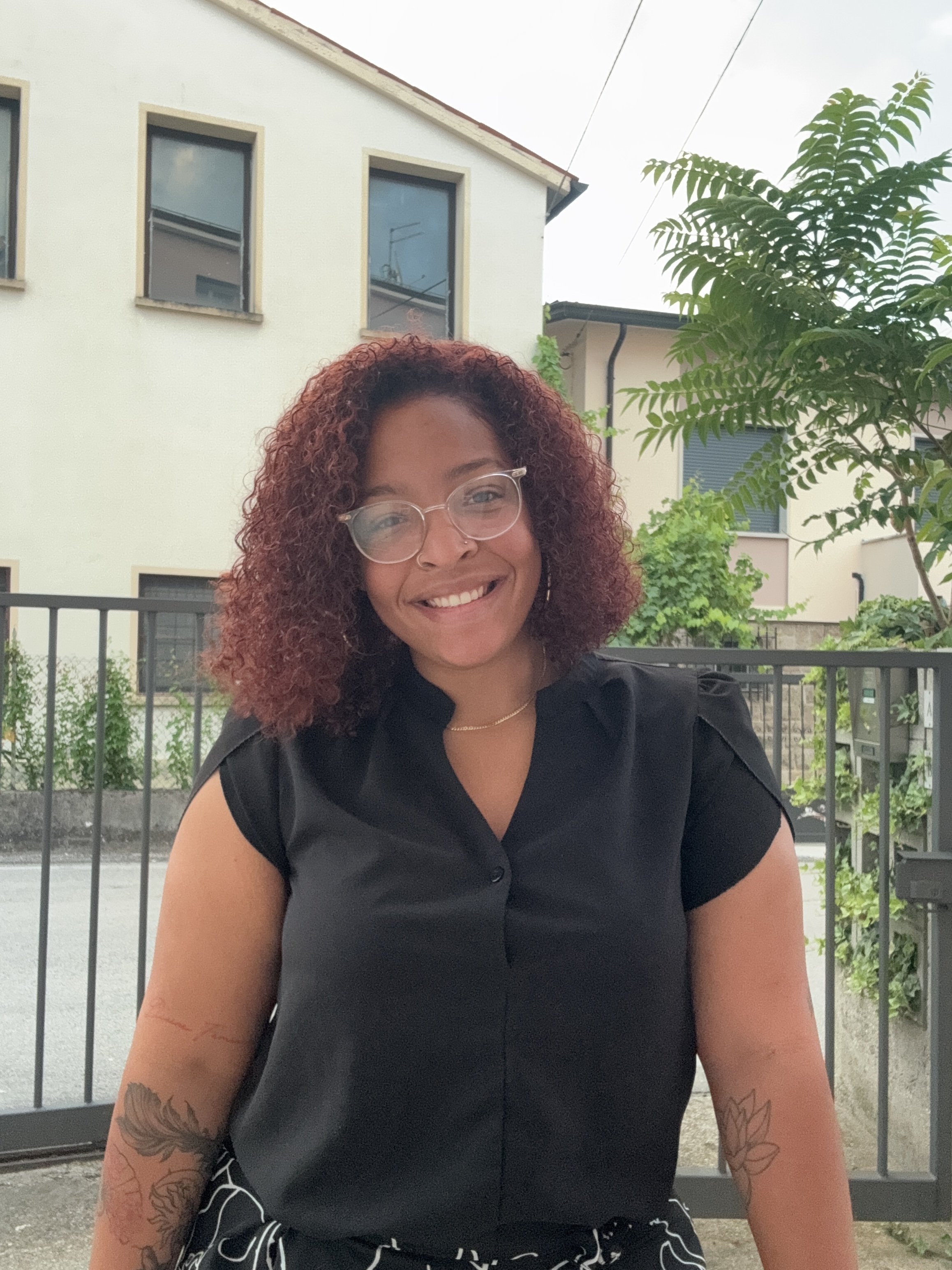 A smiling woman with curly reddish hair, glasses, and tattoos on her arms, sitting outdoors in a residential area with trees and buildings in the background.