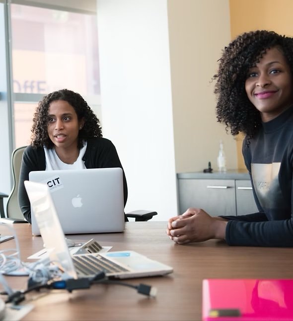 Two women sitting at a conference table in a meeting room, one with a silver MacBook laptop, engaged in conversation.