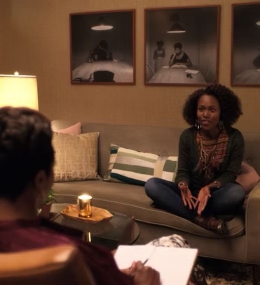 Two women having a conversation in a cozy living room, one sitting on a sofa and the other facing her with a notepad, with framed photos hanging on the wall behind.
