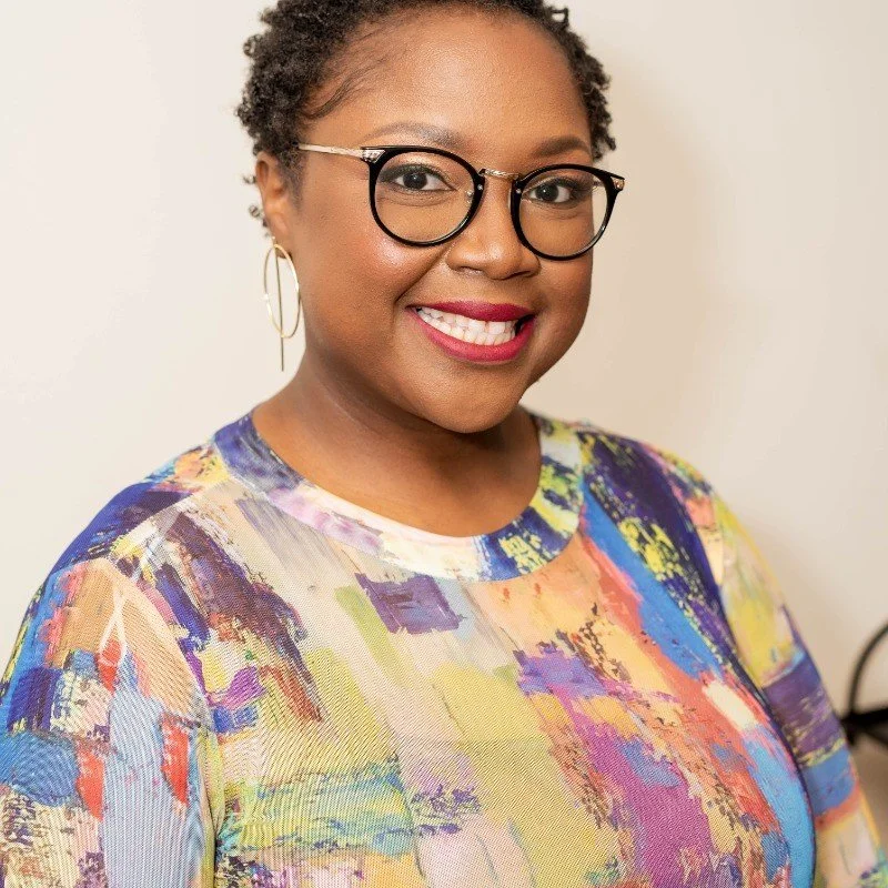 A smiling woman with short, curly hair, glasses, and hoop earrings, wearing a colorful, abstract-patterned top.