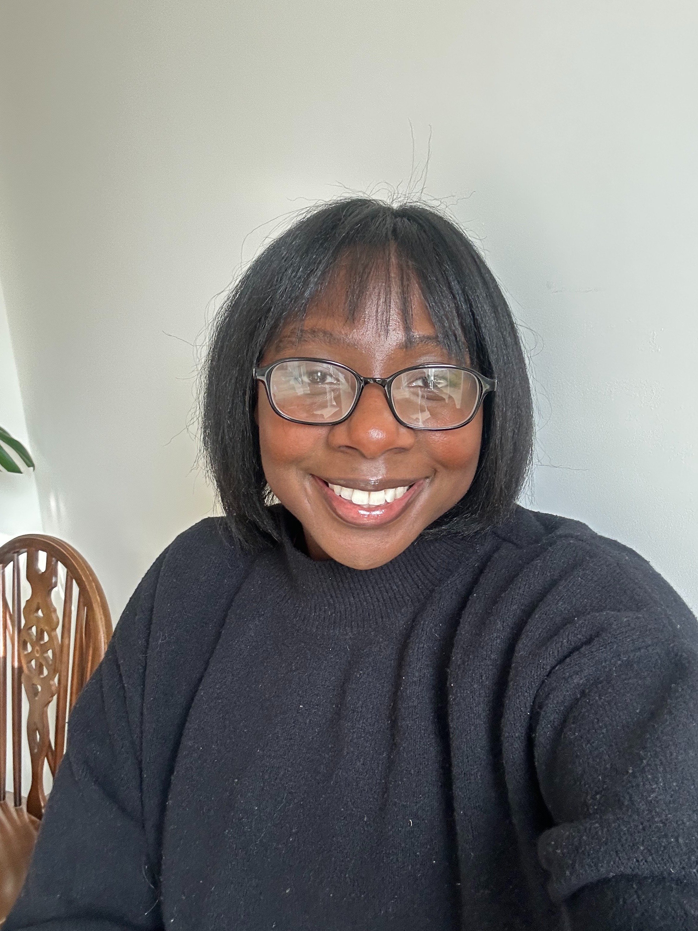 A woman with black hair, glasses, and a bright smile taking a selfie indoors in front of a white wall, with a wooden chair visible in the background.