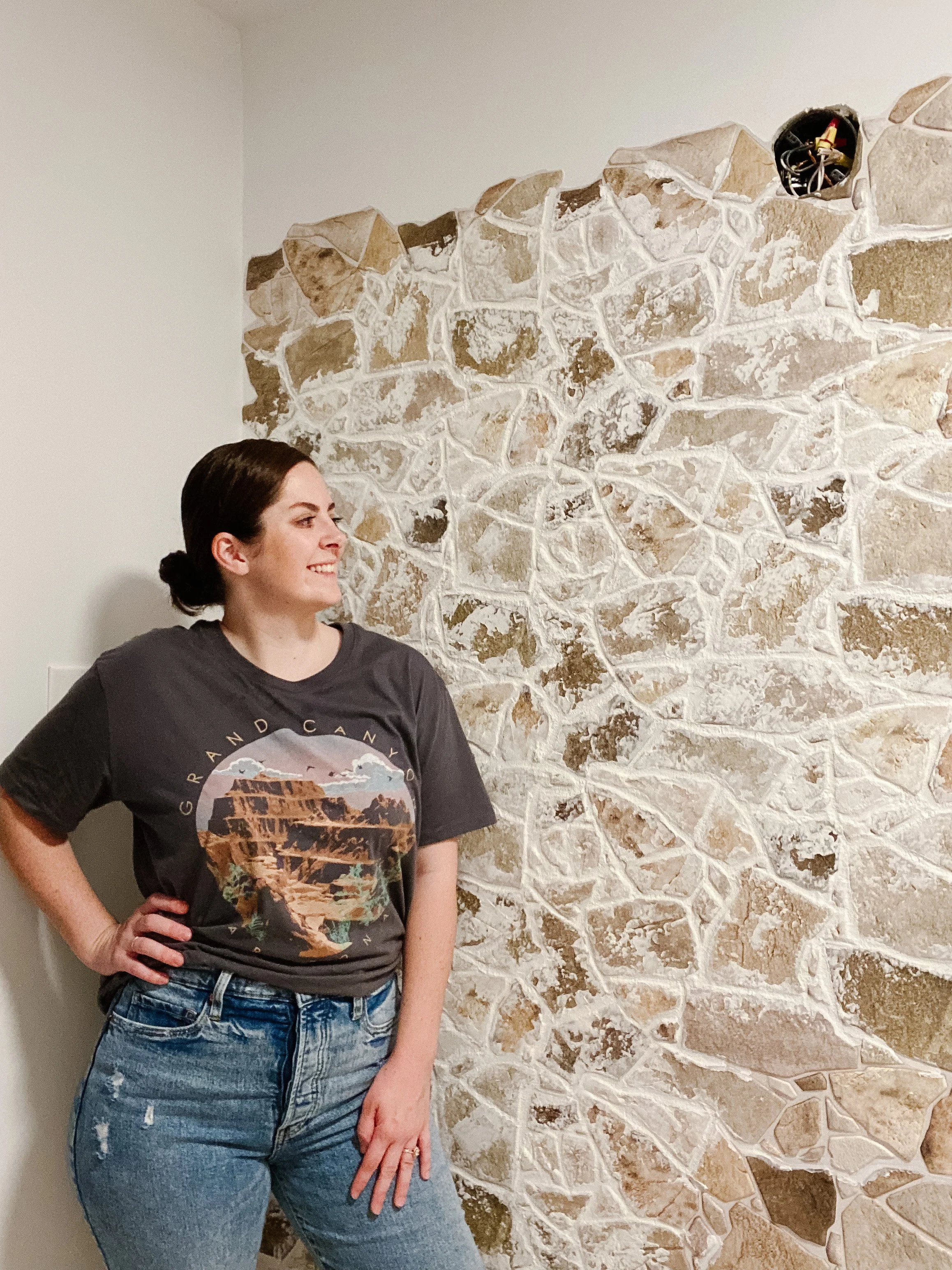 A woman with dark hair tied back, wearing a graphic t-shirt and jeans, stands next to a stone wall with an electrical outlet and wires near the top of the wall.