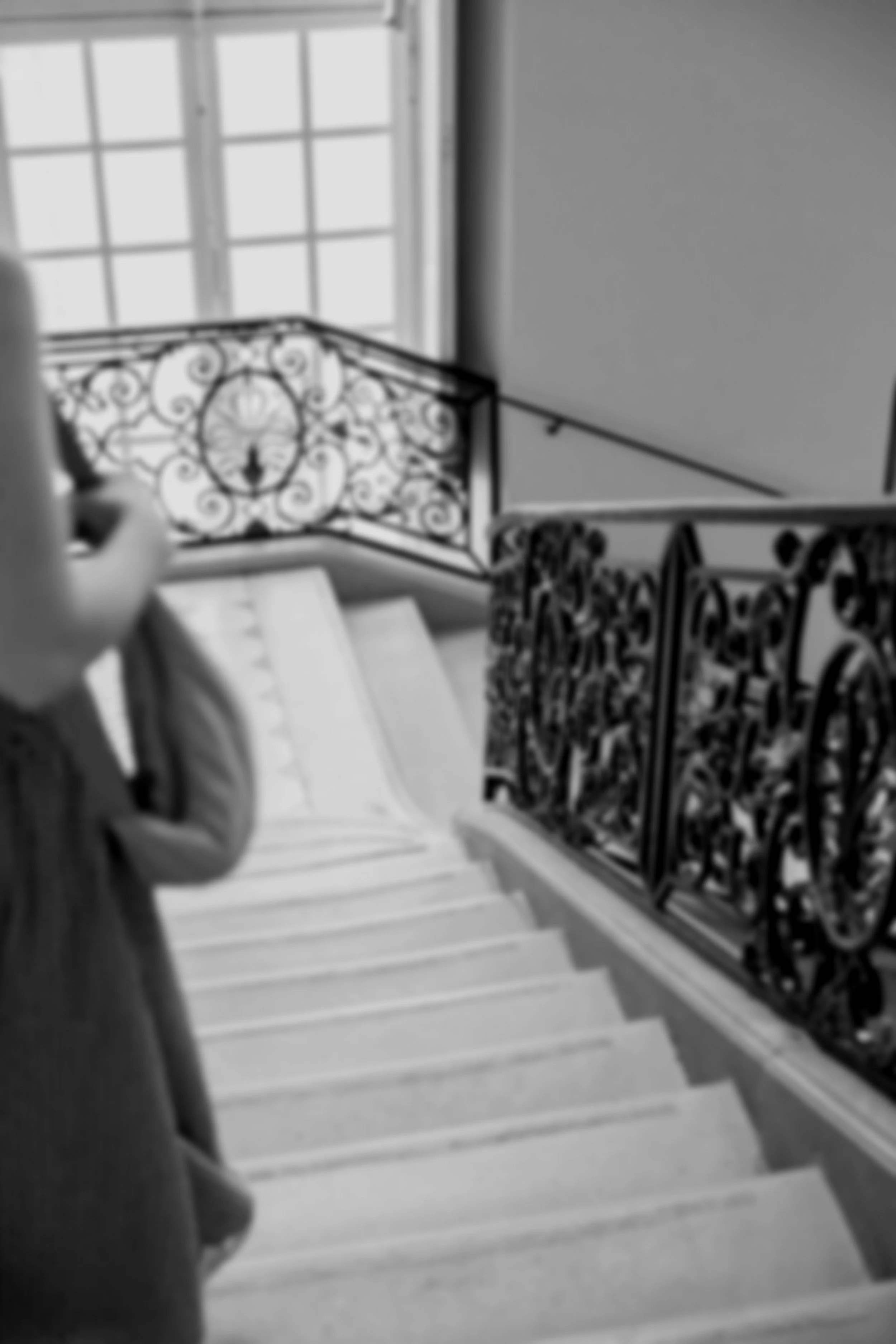A black and white photo of a grand, Parisian staircase. The stairs are wide and there's an ornate banister running alongside. In the bottom right corner, a woman starts to appear to descend the steps while still half out of shot.