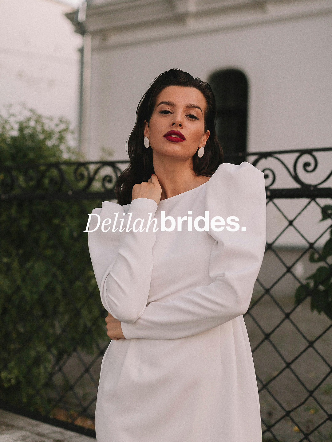 The "Delilah Brides" logo (the name written in all white text with Delilah in a serif font and brides in a sans-serif), sits on top of an image of woman in white dress. The dress is modern, shift-like dress with long sleeves.