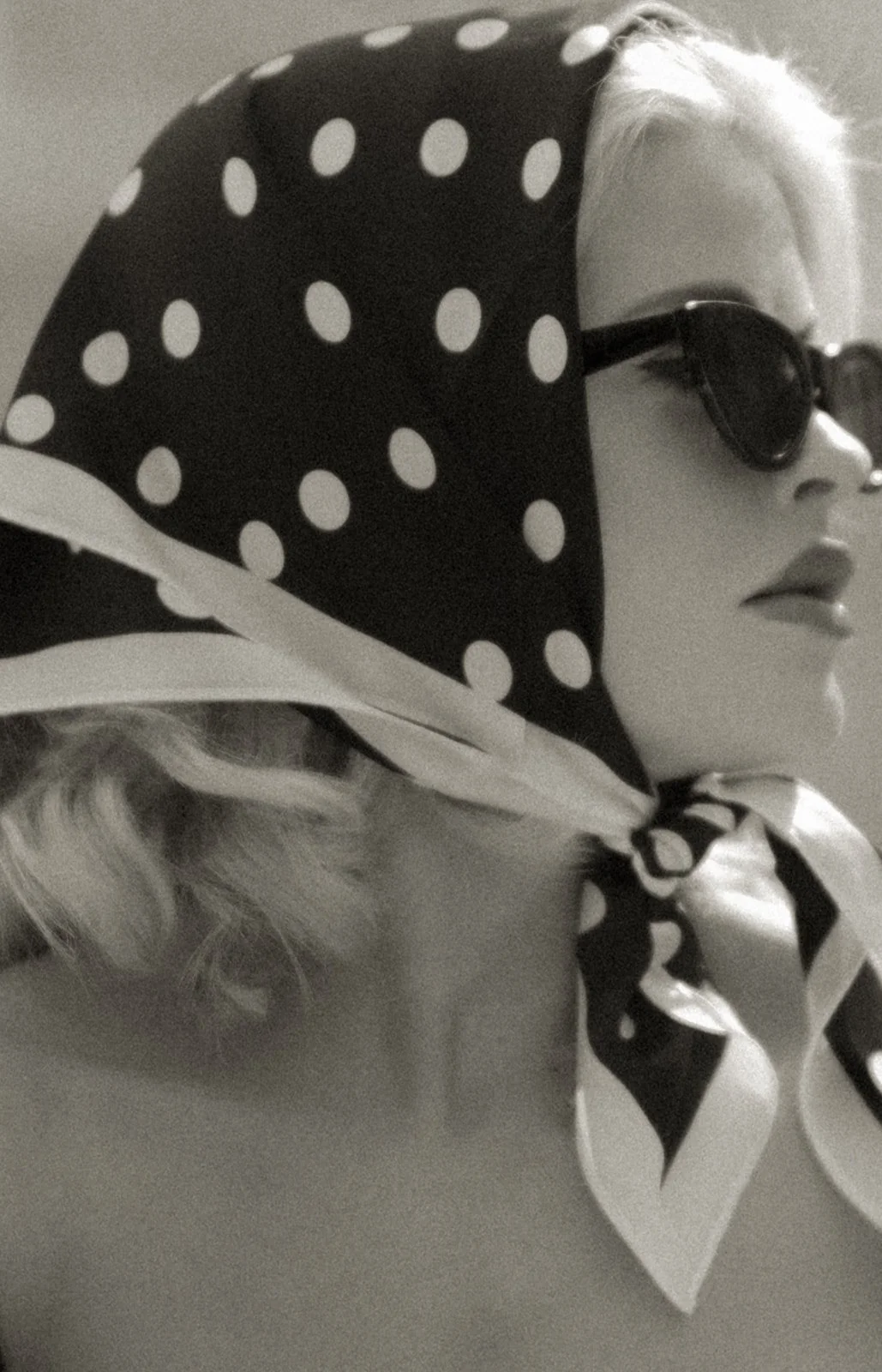 A woman looks off in the distance wearing a dark headscarf with white trims and polka dots. She's also wearing sunglasses, depicting a 1950s style era