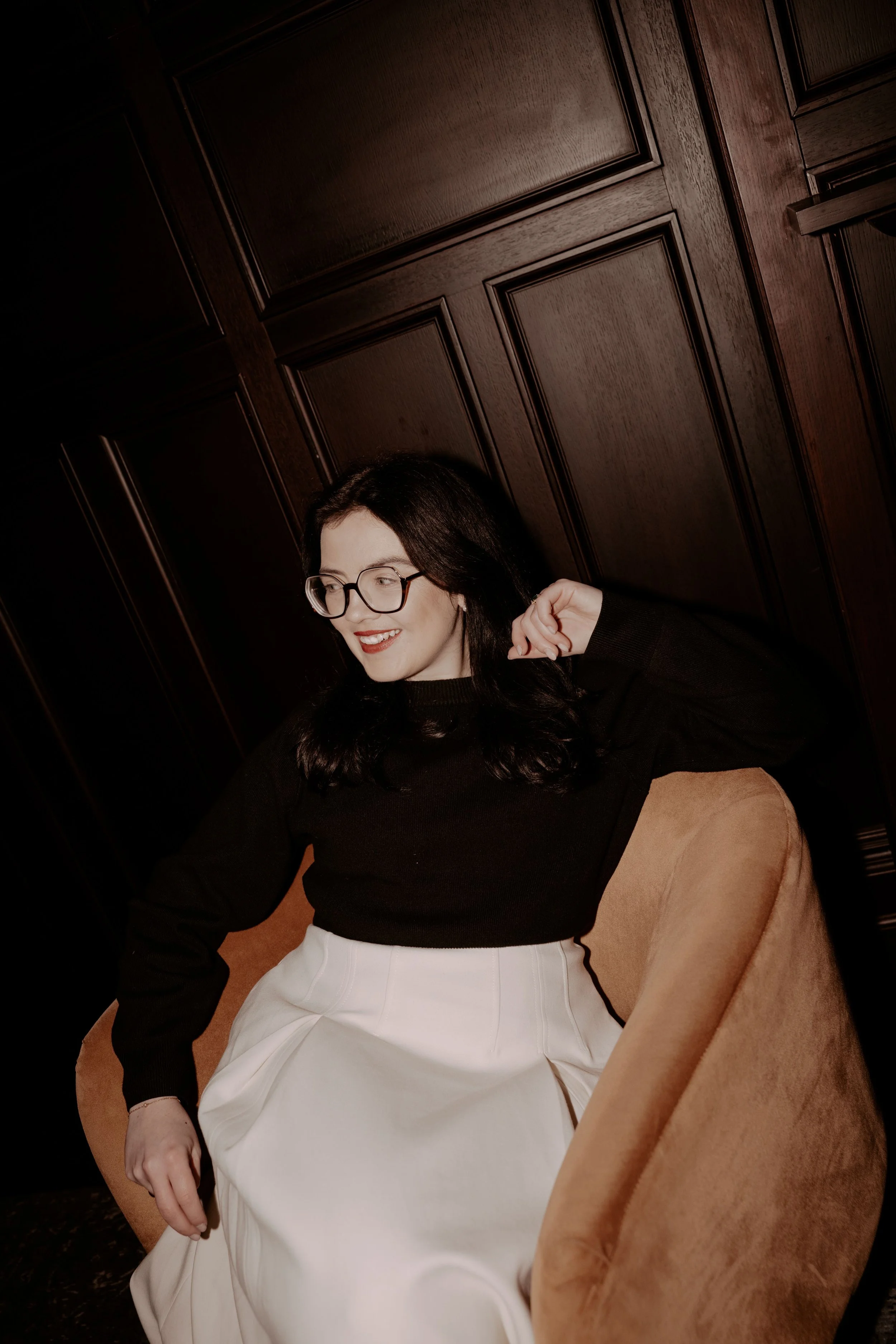 Soph is sat on a chair in a black jumper and white skirt. She's wearing glasses and red lipstick and smiling + looking confident into the distance. Behind her is a dark wood panelled wall.