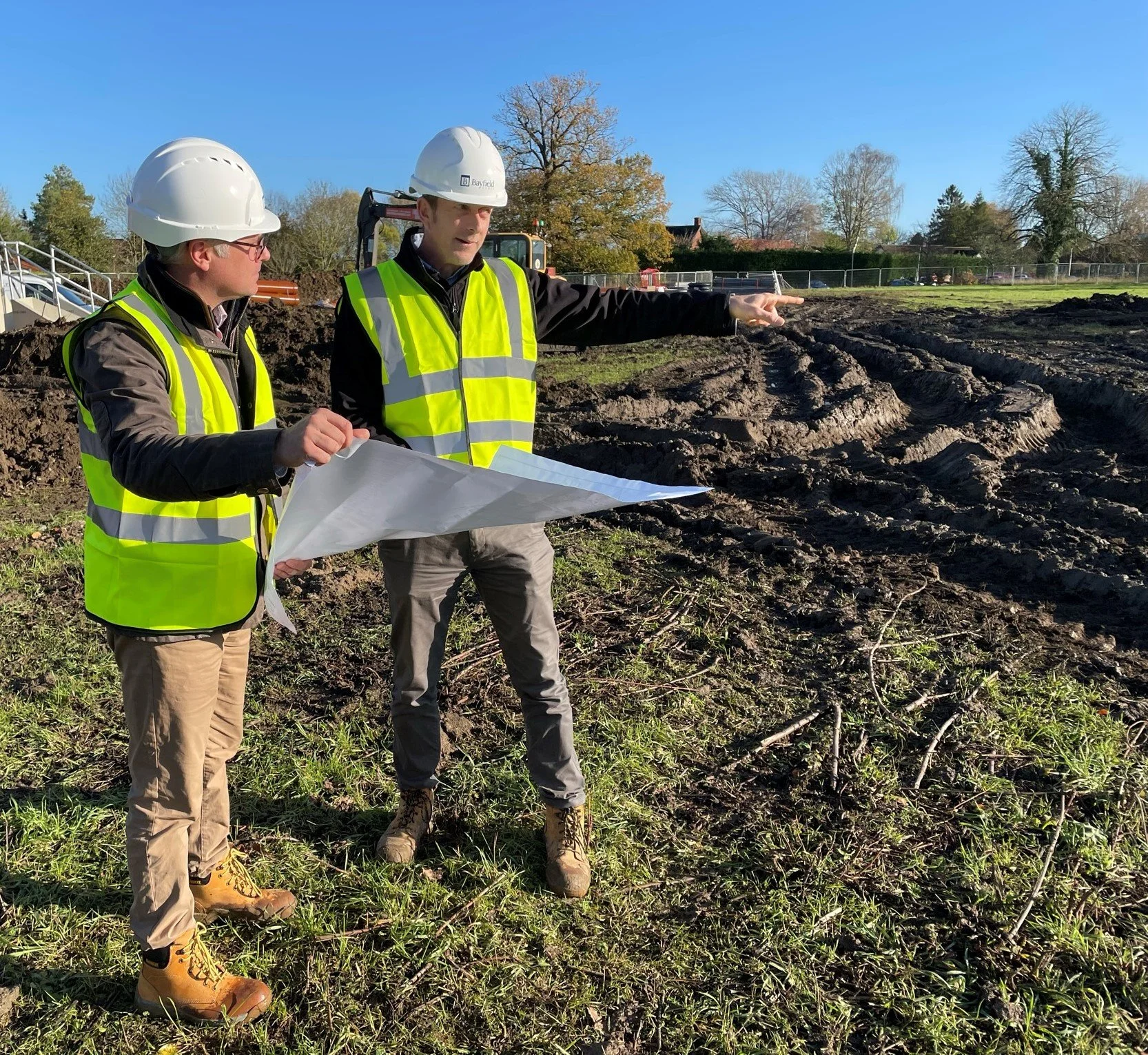 Two construction workers in high-visibility vests and helmets at a construction site, examining blueprints and pointing at the ground.