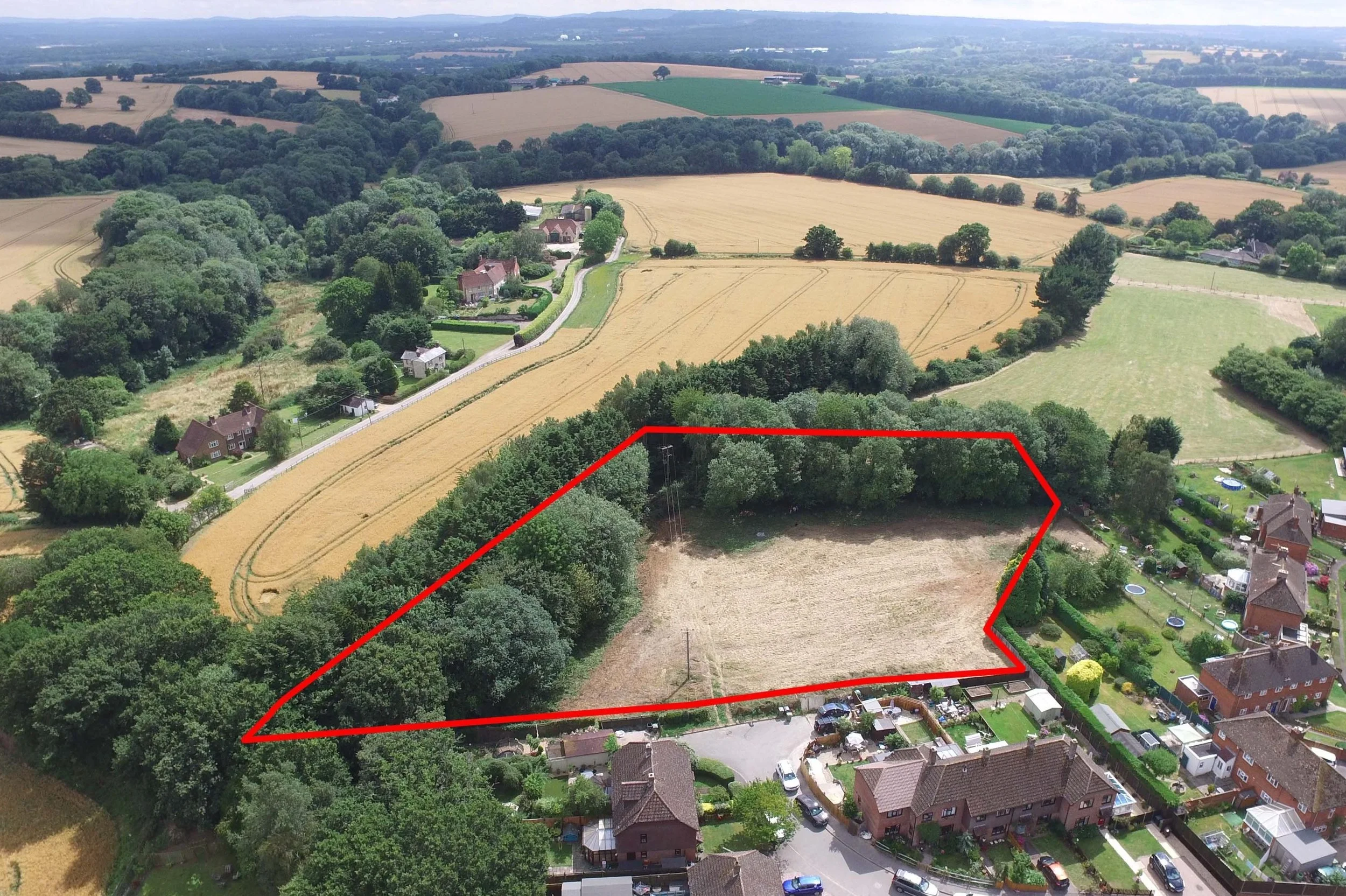 Aerial view of a rural landscape with a triangular plot of land outlined in red, surrounded by trees and near residential houses and fields.
