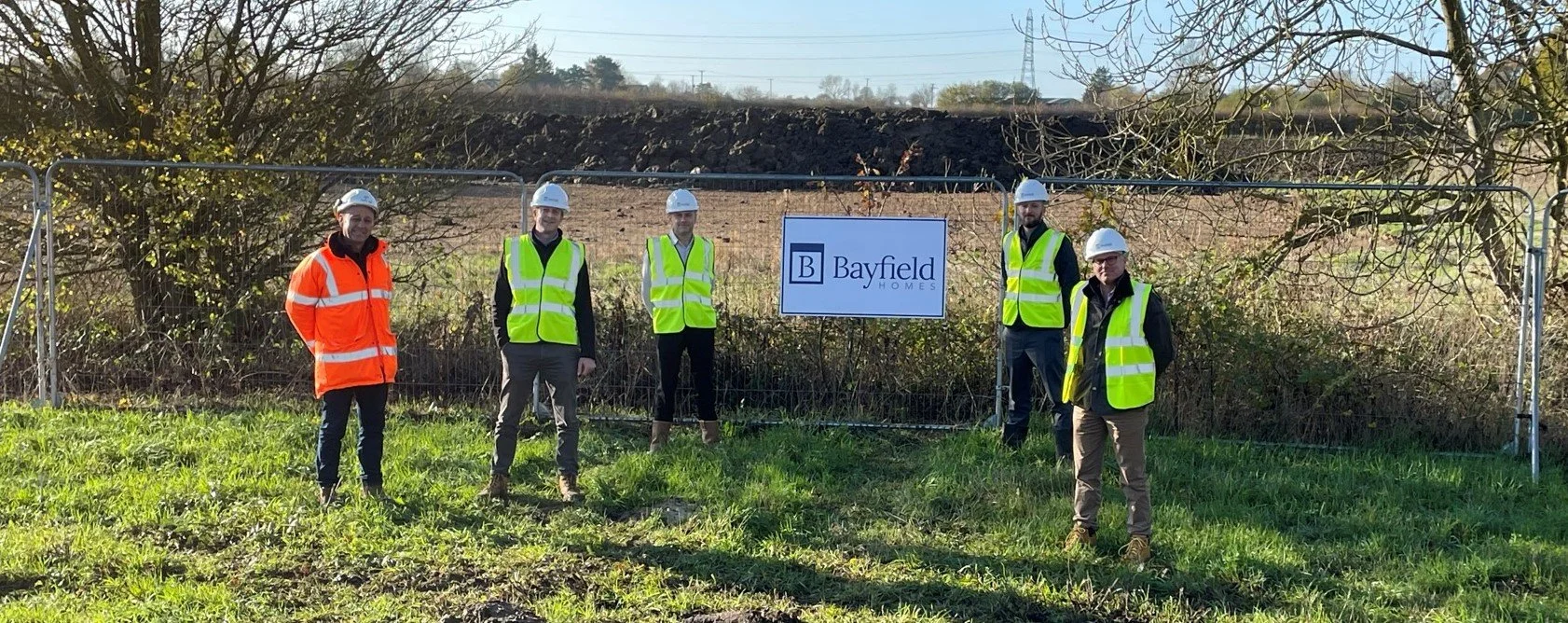 Group of five people in construction gear, including hard hats and high-visibility vests, standing outdoors in front of a "Bayfield Homes" sign and a metal fence on a grassy area.