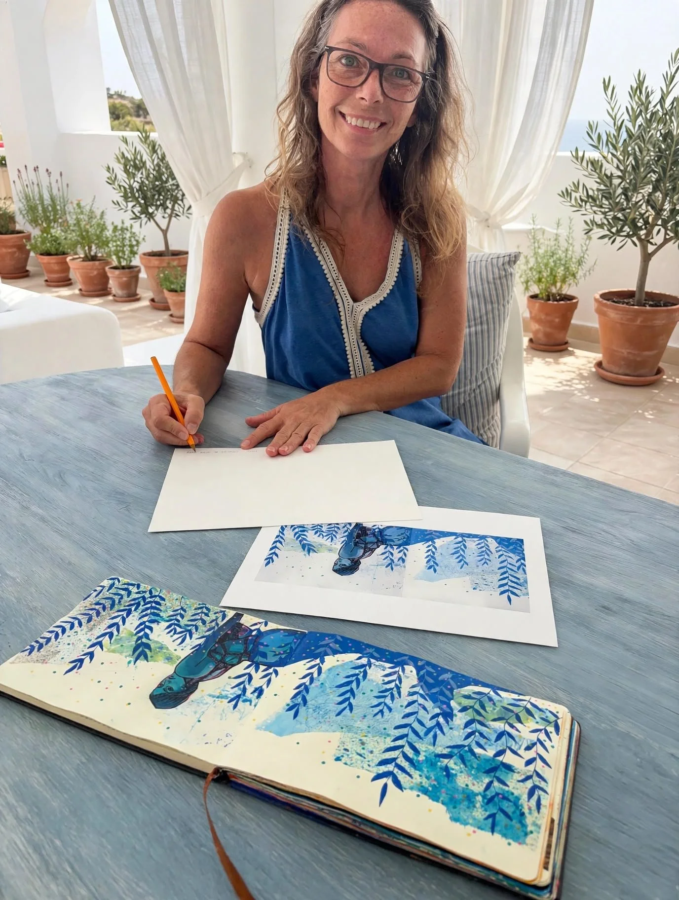 Mujer sonriendo, sentada en una mesa de madera gris, firmando un papel con un bolígrafo naranja, en un entorno con muchas plantas en macetas de barro y cortinas blancas, en un lugar con vista al mar.