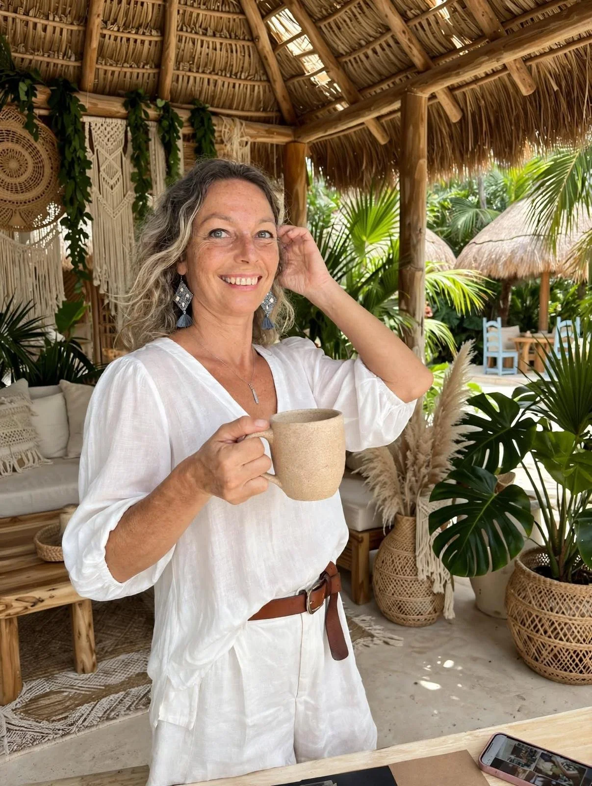 Mujer joven con cabello rizado , vestida con blusa blanca, sosteniendo una taza en un ambiente de estilo tropical, rodeada de plantas y decoraciones de mimbre en un espacio abierto con techo de hojas de palma.
