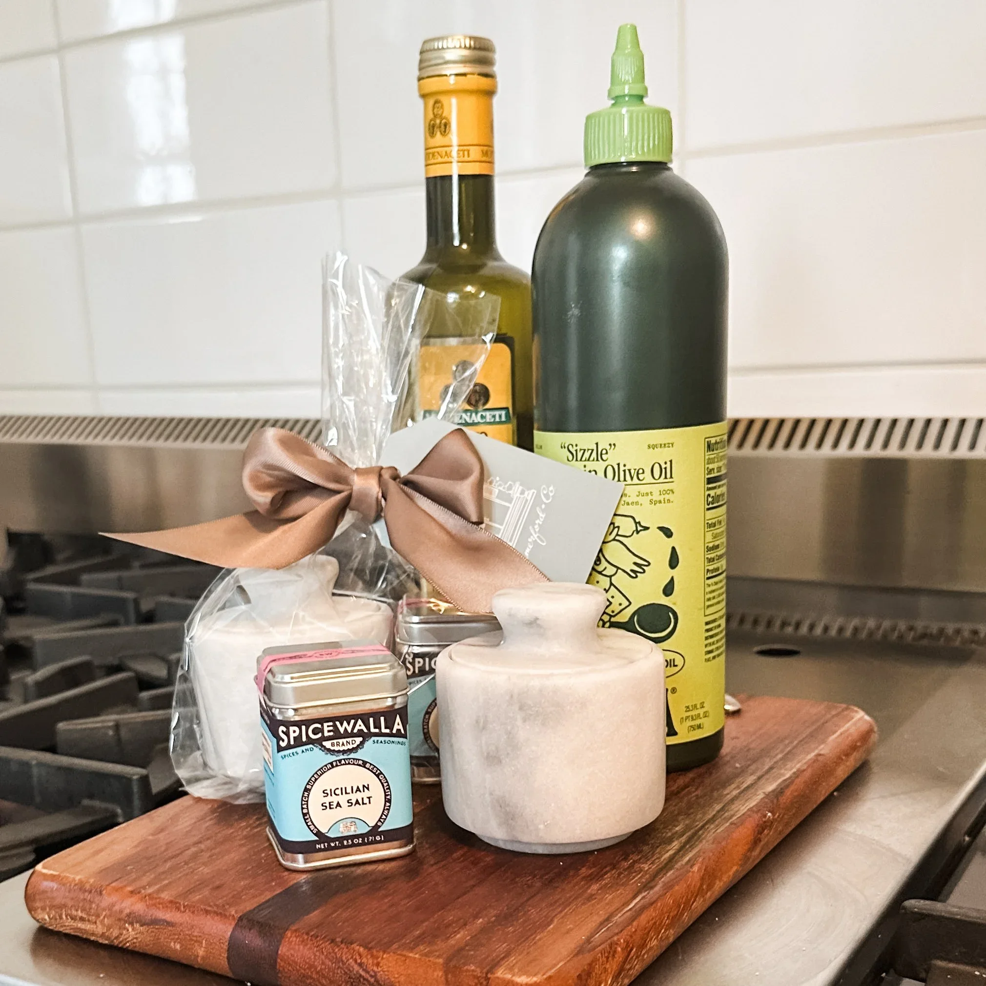 A bottle of Sicilian sea salt, a container of Sizzle Olive Oil, and a small pink container on a wooden cutting board in a kitchen setting.