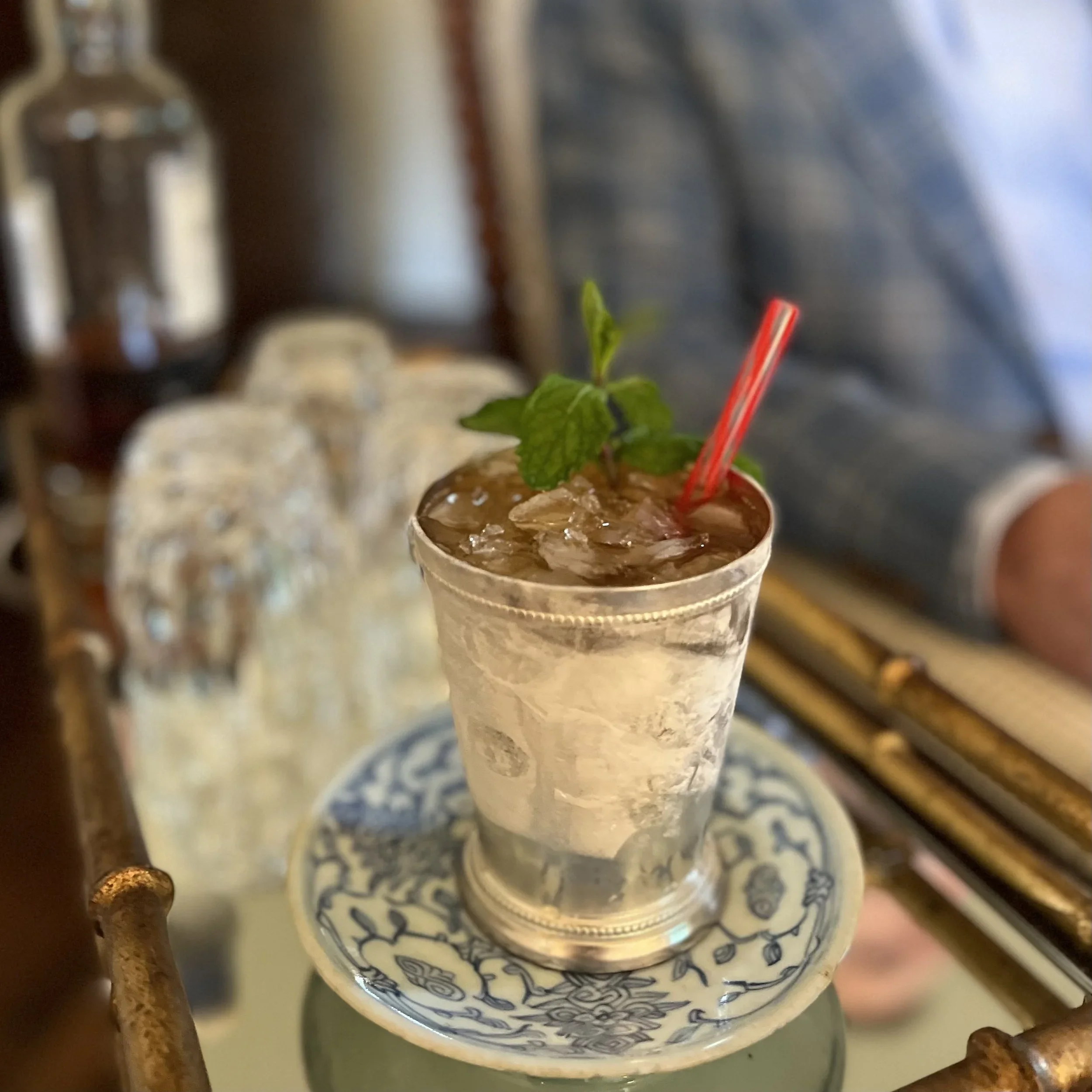 A close-up of a blue and white patterned ceramic cup filled with a beverage topped with crushed ice, garnished with a sprig of mint and a red straw, with a blurred background of a bar or café setting with bottles and glassware.