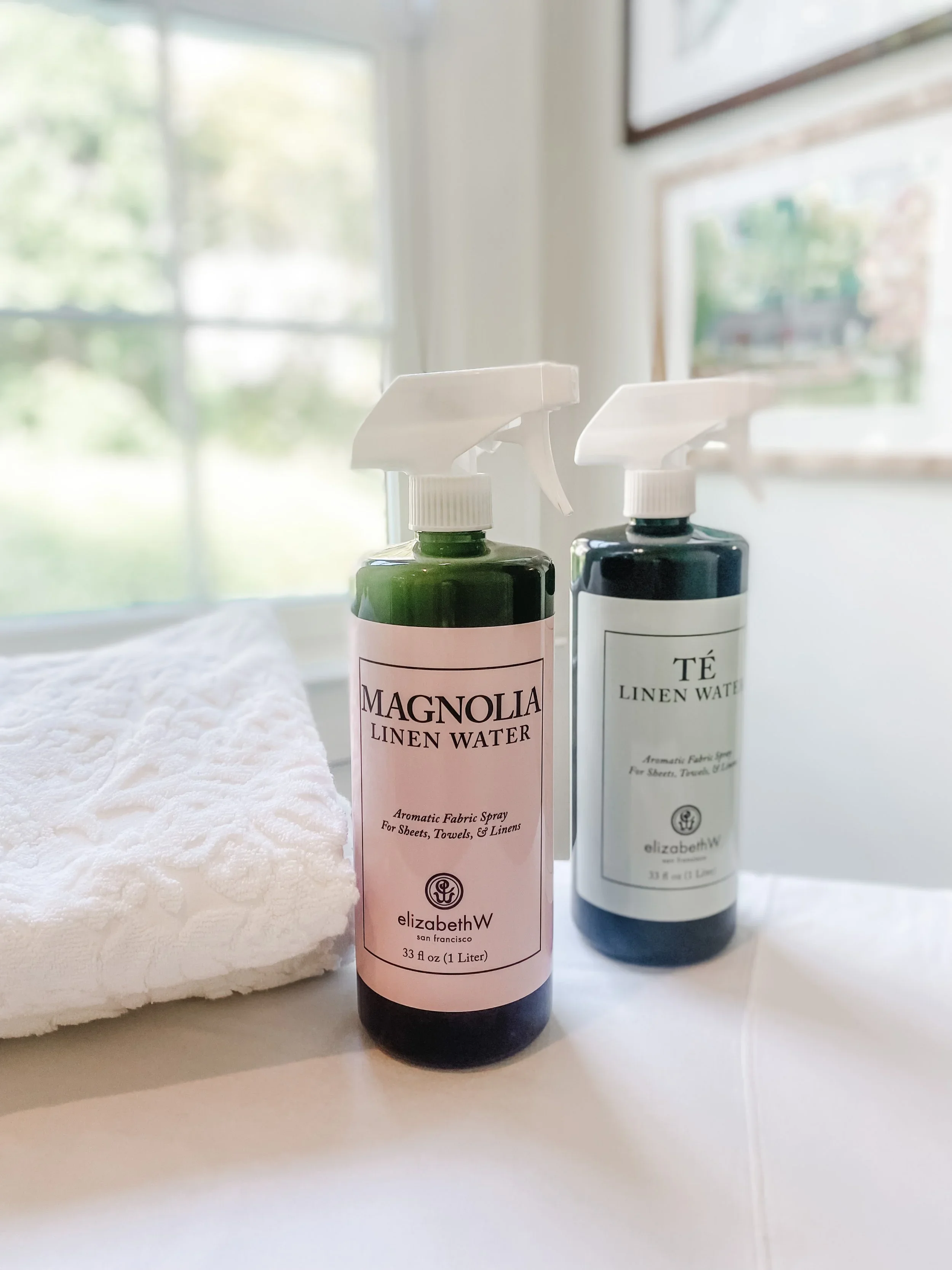 Two bottles of linen water spray, one pink labeled 'Magnolia Linen Water' and one white labeled 'Té Linen Water', placed on a white surface next to a white folded towel, with a window and a blurred outdoor scene in the background.