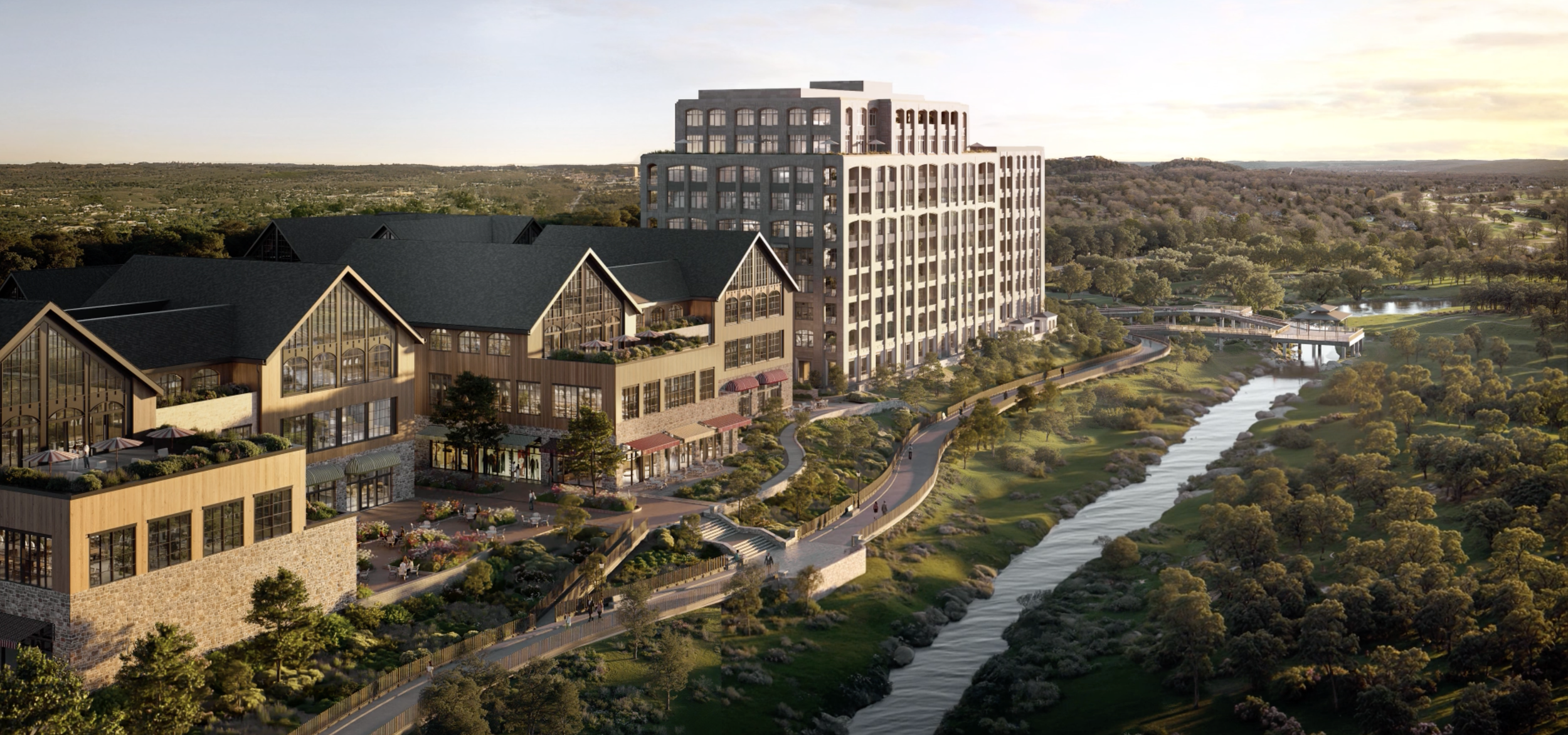 A large multi-story building complex with a mix of modern and rustic architecture situated along a river in a lush, green landscape during sunset.