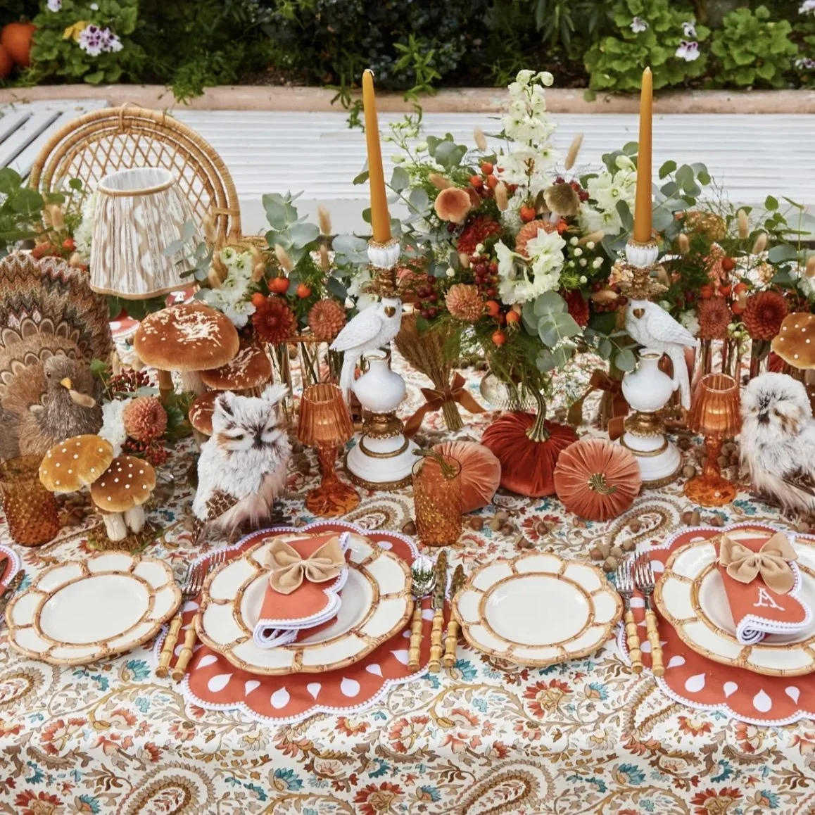 Table setup for a fall or autumn-themed gathering with floral arrangements, plush pumpkins, mushrooms, owls, candles, and decorative plates and napkins, all in warm earthy tones.