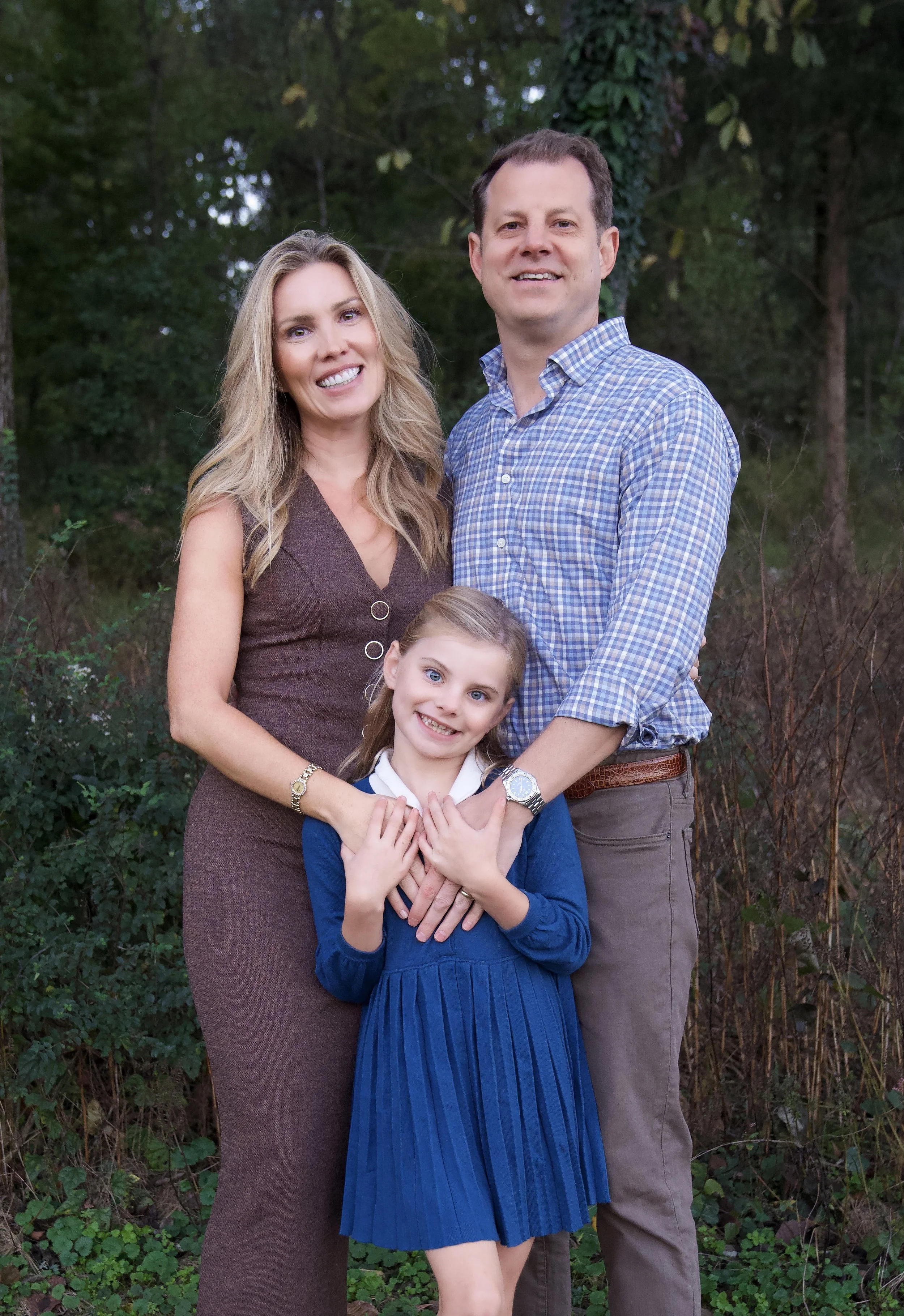 A smiling family of three, including a woman, a man, and a young girl, standing outdoors in a wooded area.