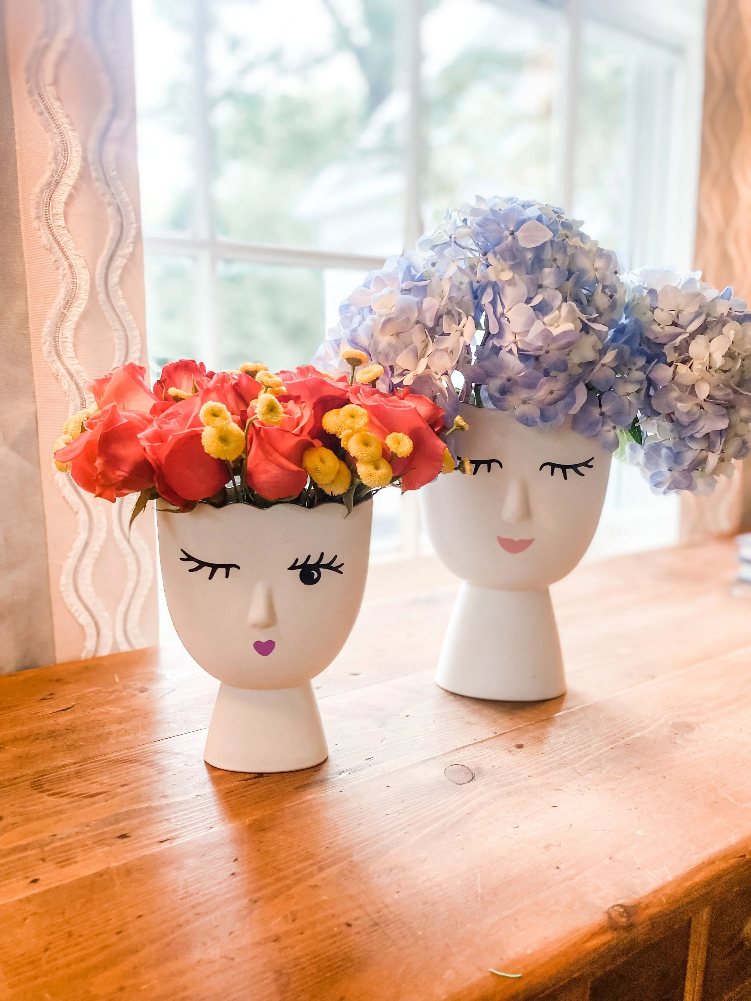 Two decorative flower vases with face designs, one with blue hydrangeas and the other with pink and yellow flowers, placed on a wooden table near a window.