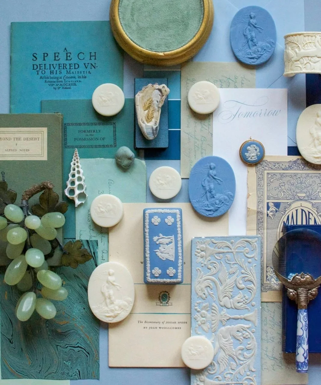 A collection of vintage and decorative items, including books, soap with embossed figures, carved stones, a small sculpture, a bunch of grapes, and ornate papers and stationery, arranged in a collage-style display with a blue and cream color theme.