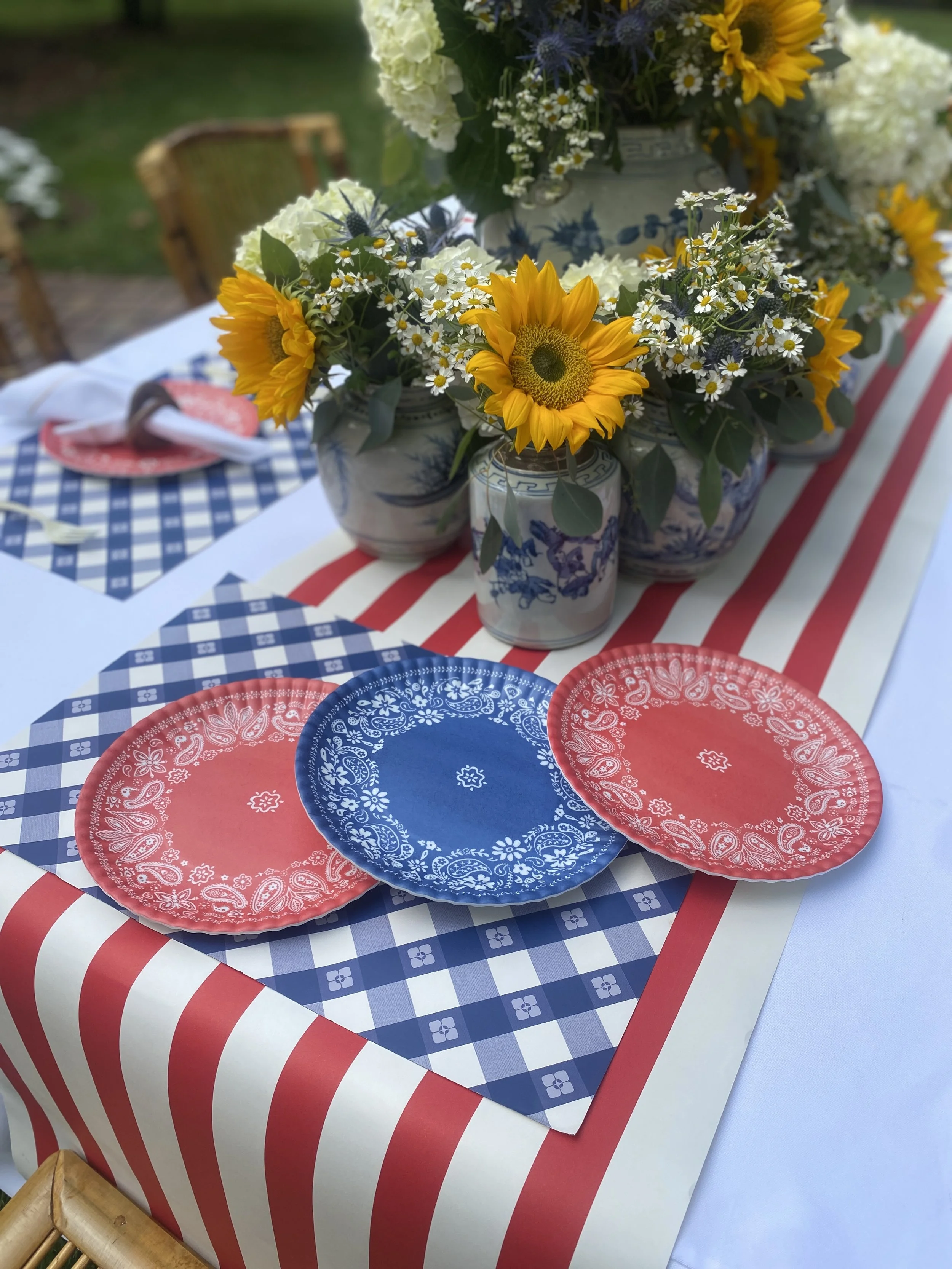 Table decorated with a blue and white checkered tablecloth, with red and blue paper plates, and a centerpiece of sunflowers and daisies in ceramic vases, outdoors.
