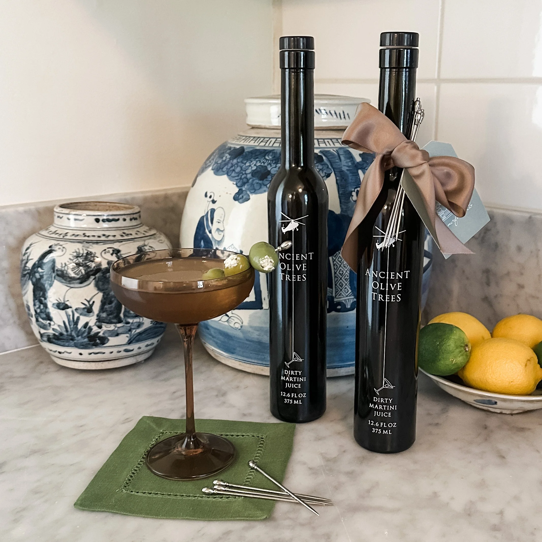 A decorative ceramic vase with blue floral designs, a small round wooden cocktail glass with green olives, a green placemat, and two bottles of Ancient Olive Trees dirty martini juice on a white marble countertop.