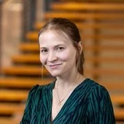 A young woman with reddish-brown hair smiling in front of a wooden wall.