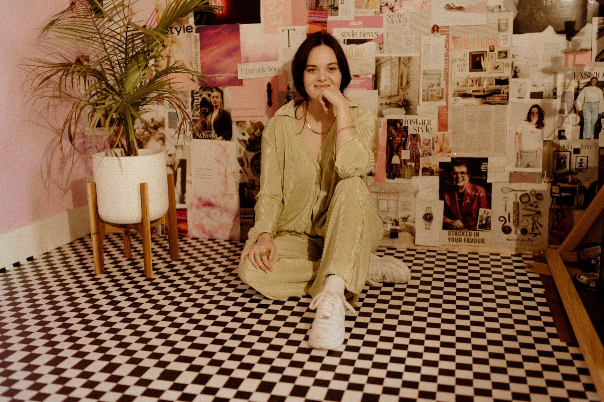A young woman with dark hair sitting on the floor in a room with a pink wall and a collage of magazine cutouts and posters on the wall behind her. She is wearing a yellow outfit, white sneakers, and has her hand resting on her chin, smiling slightly. To her left, there is a potted plant on a wooden stand with a black-and-white checkered floor.