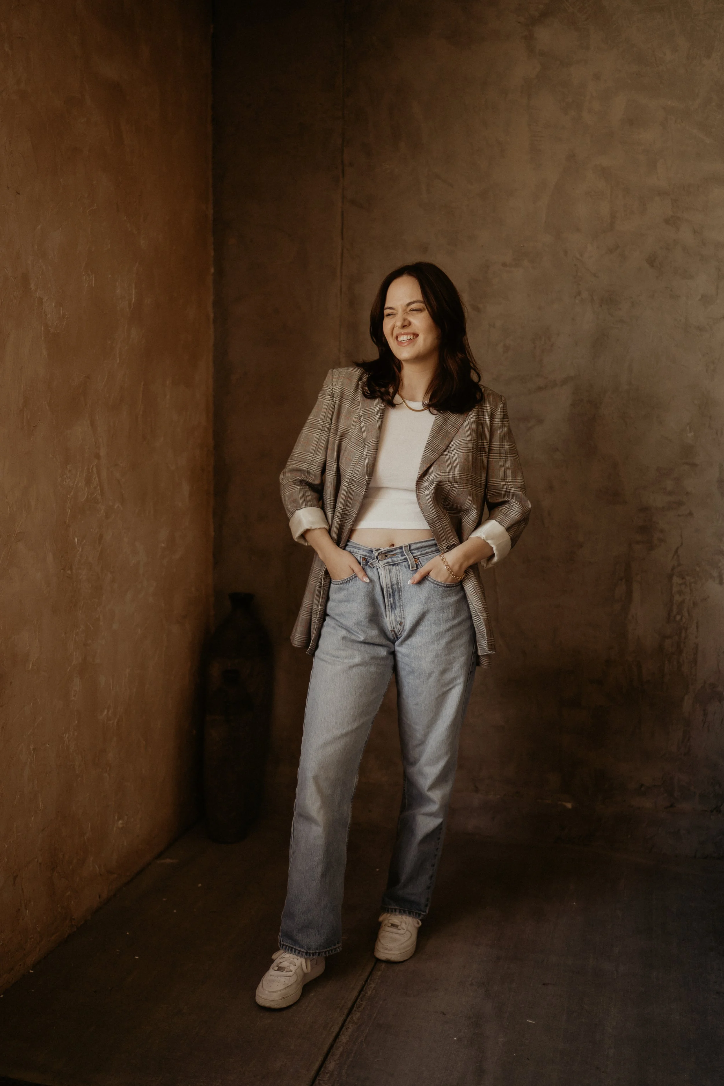 A young woman with dark hair wearing a plaid blazer, white crop top, and baggy jeans standing in a dimly lit space with textured brown walls, smiling with her eyes closed.