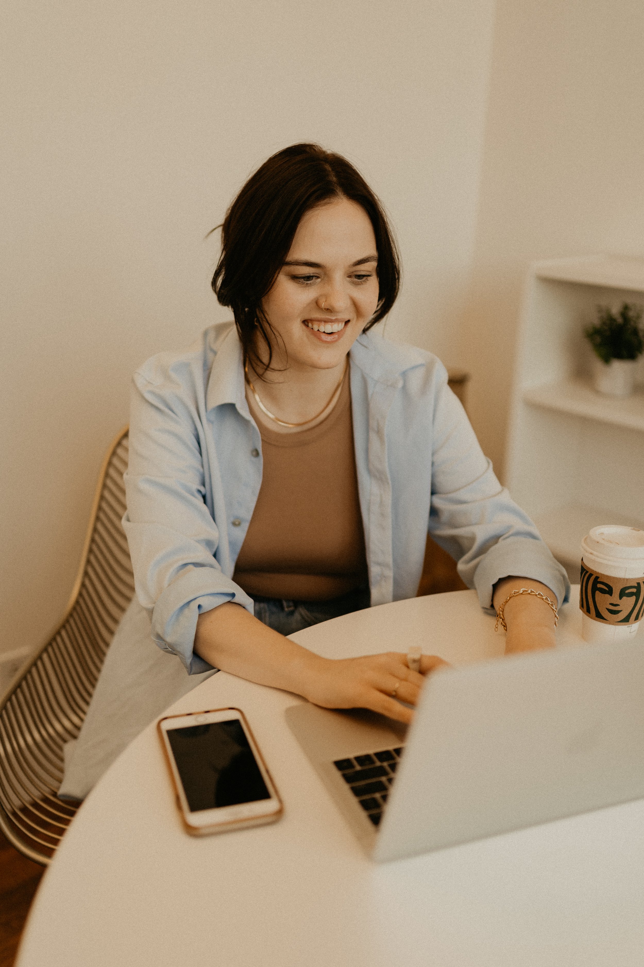 A young woman with dark hair and a nose ring sitting at a white table, smiling and looking down. On the table, there is a smartphone, an open laptop, and a coffee cup, with a smartphone case and a laptop in the foreground.