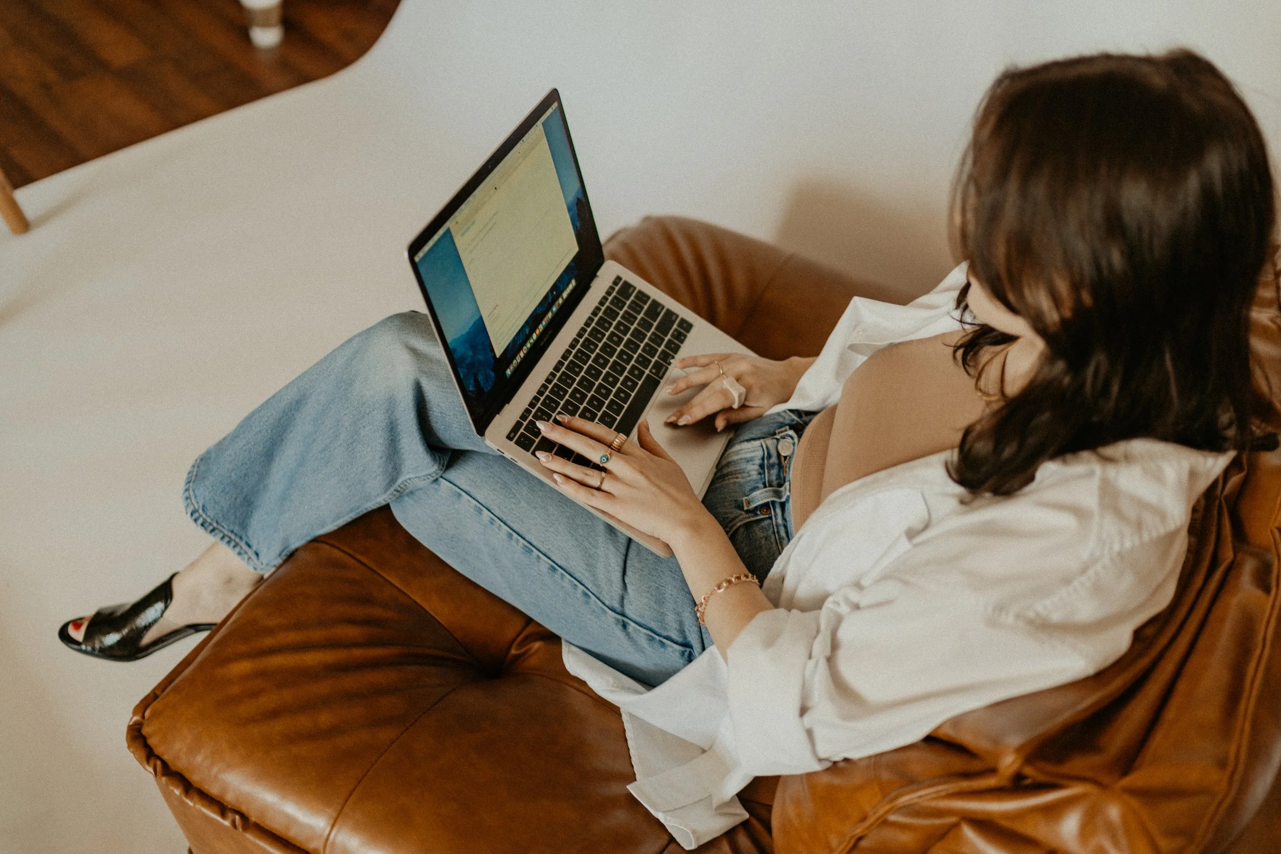A woman sitting on a brown leather couch, working on a laptop, wearing casual clothes and colorful socks.
