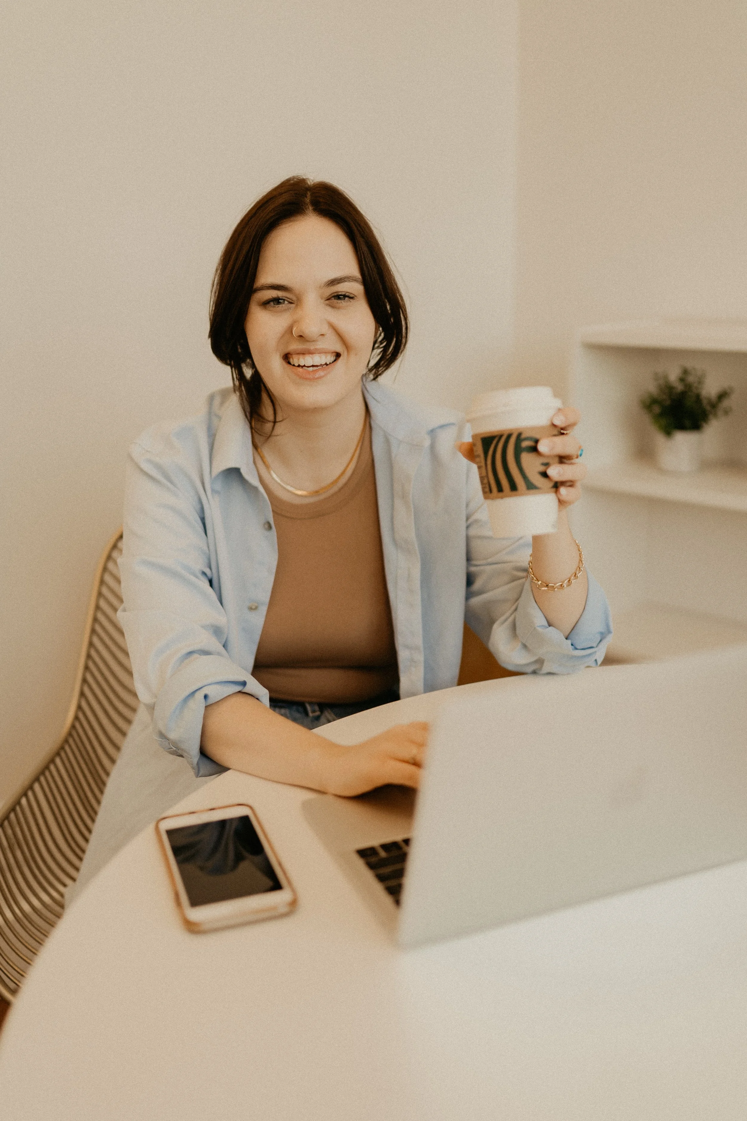 A woman with dark hair, wearing a beige top and a white shirt, smiling and holding a Starbucks coffee cup, sitting at a white table with a smartphone and a laptop on it.