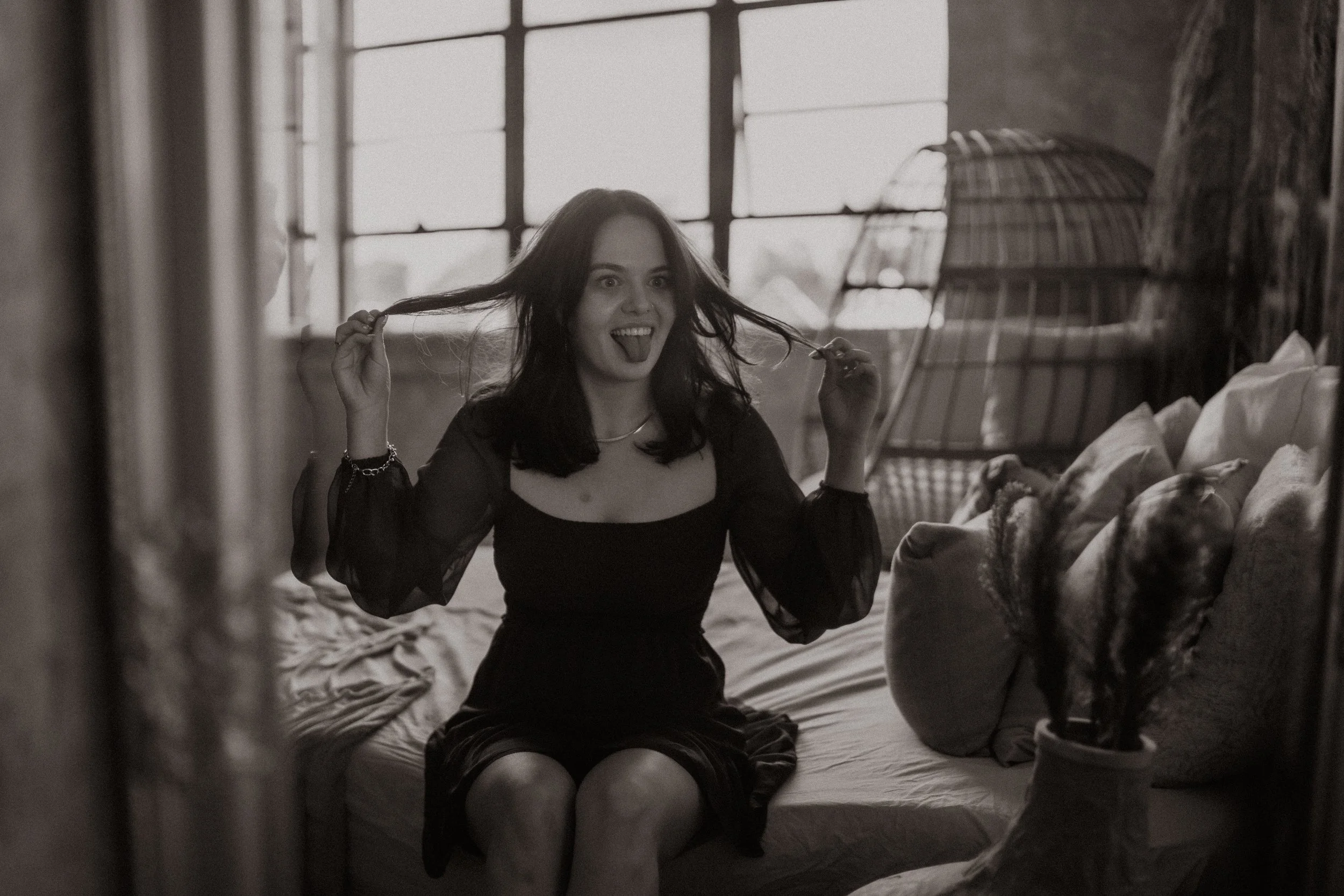A young woman with dark hair, wearing a black dress with sheer sleeves, sits on a bed in a room with large windows. She is smiling and playfully holding her hair, sticking out her tongue. The room has a rustic style with a large clay vase and bedding around her.