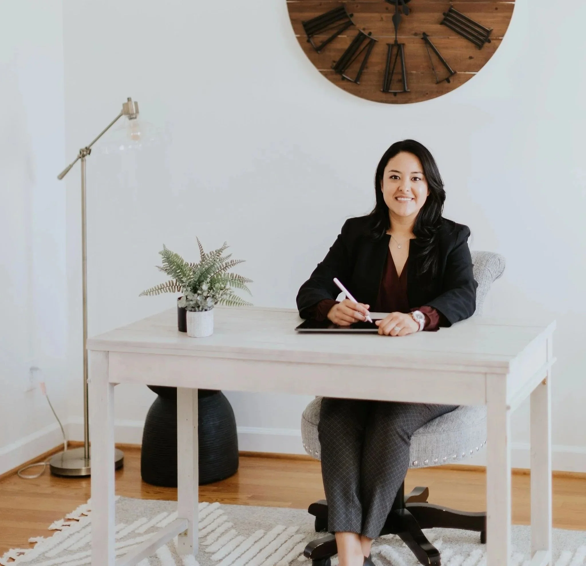 A professional Hispanic or Latina woman sitting at a white desk in a well-lit room, smiling, with a potted plant and a large wooden clock on the wall behind her.