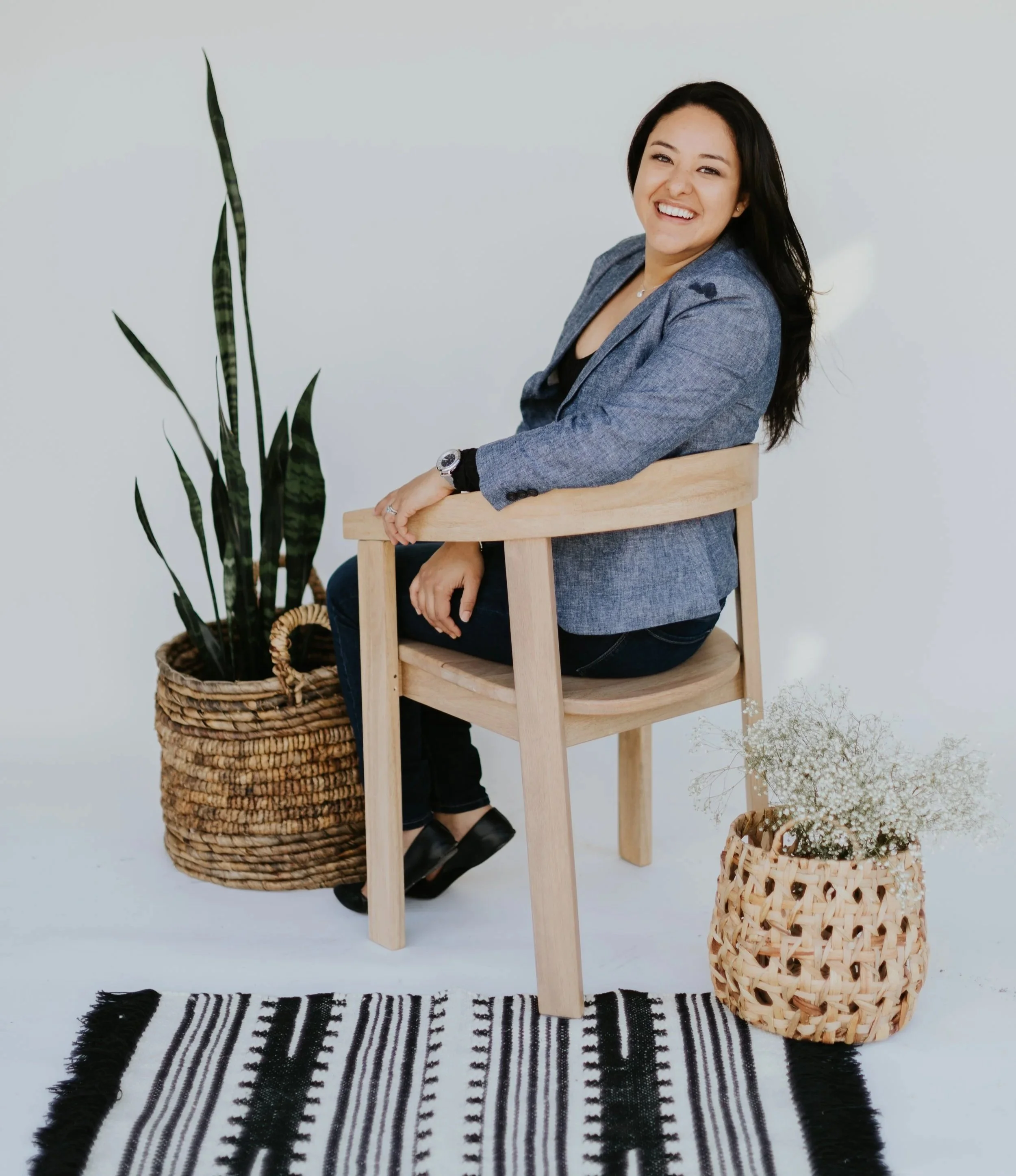 A professional hispanic and latina woman with long dark hair, smiling, sitting on a wooden chair in front of a white background, surrounded by potted plants and a patterned rug.