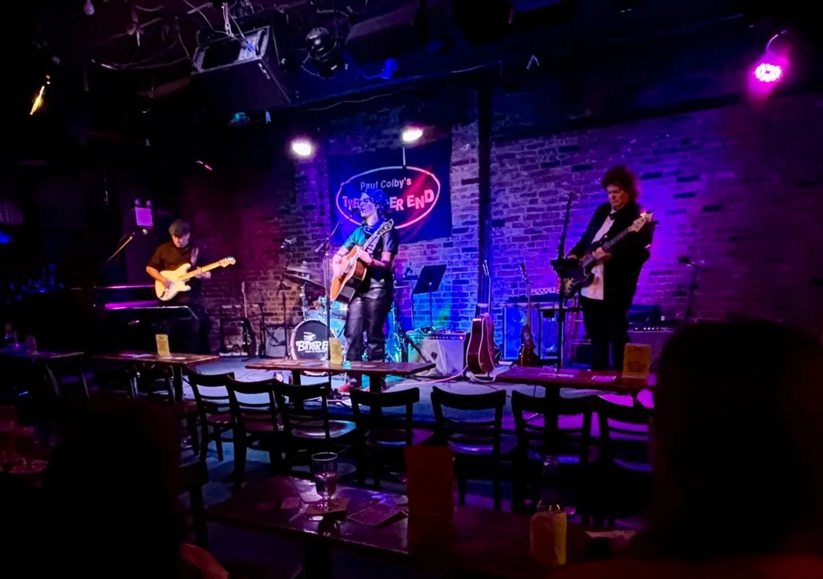 A musical band performs on a small stage in a dimly lit bar or club, with a brick wall background and a sign reading 'Paul Colby's THE END'.