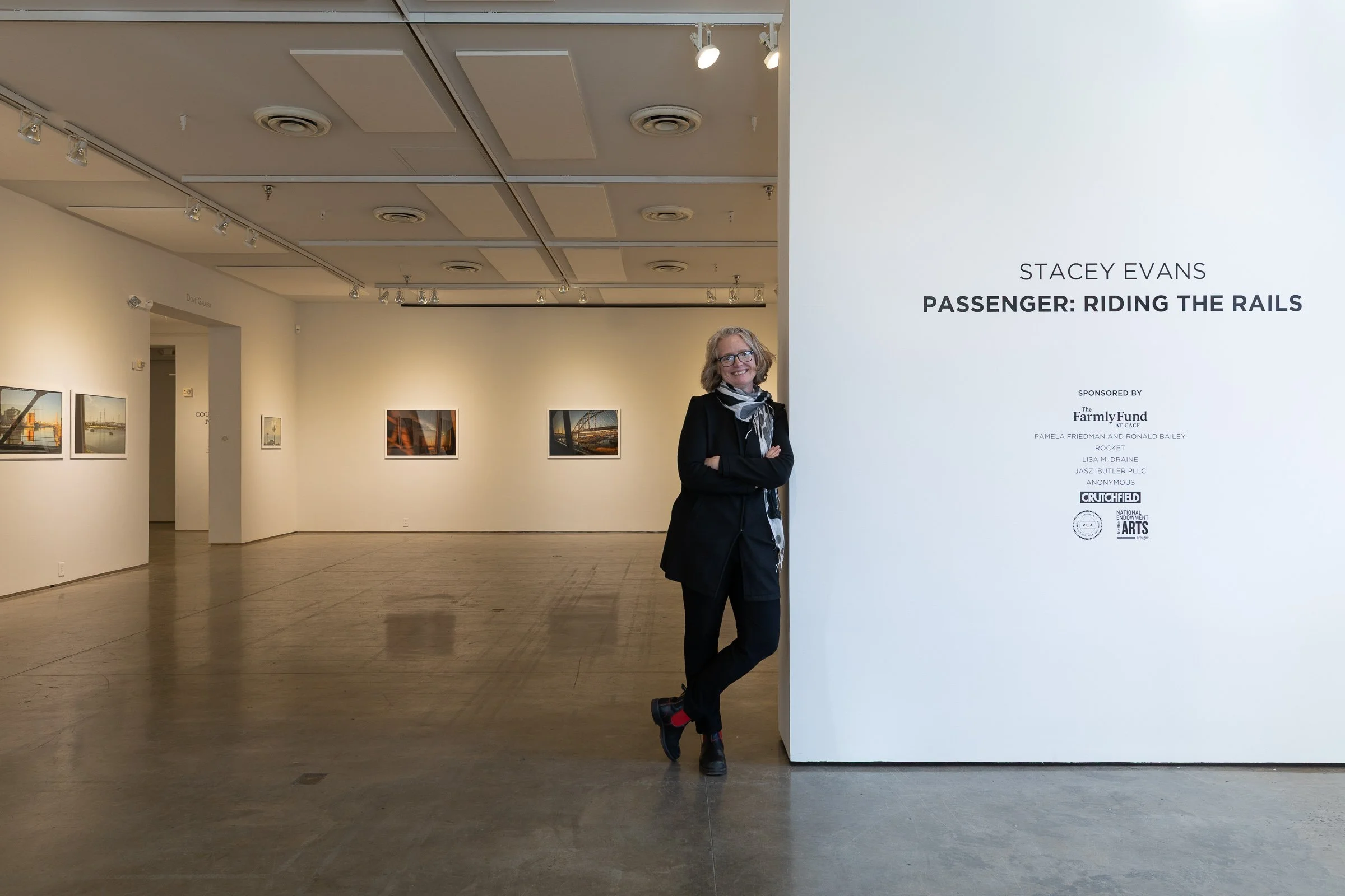 Stacey Standing in the Second Street Gallery leaning against the wall signage with framed photographs in the background.