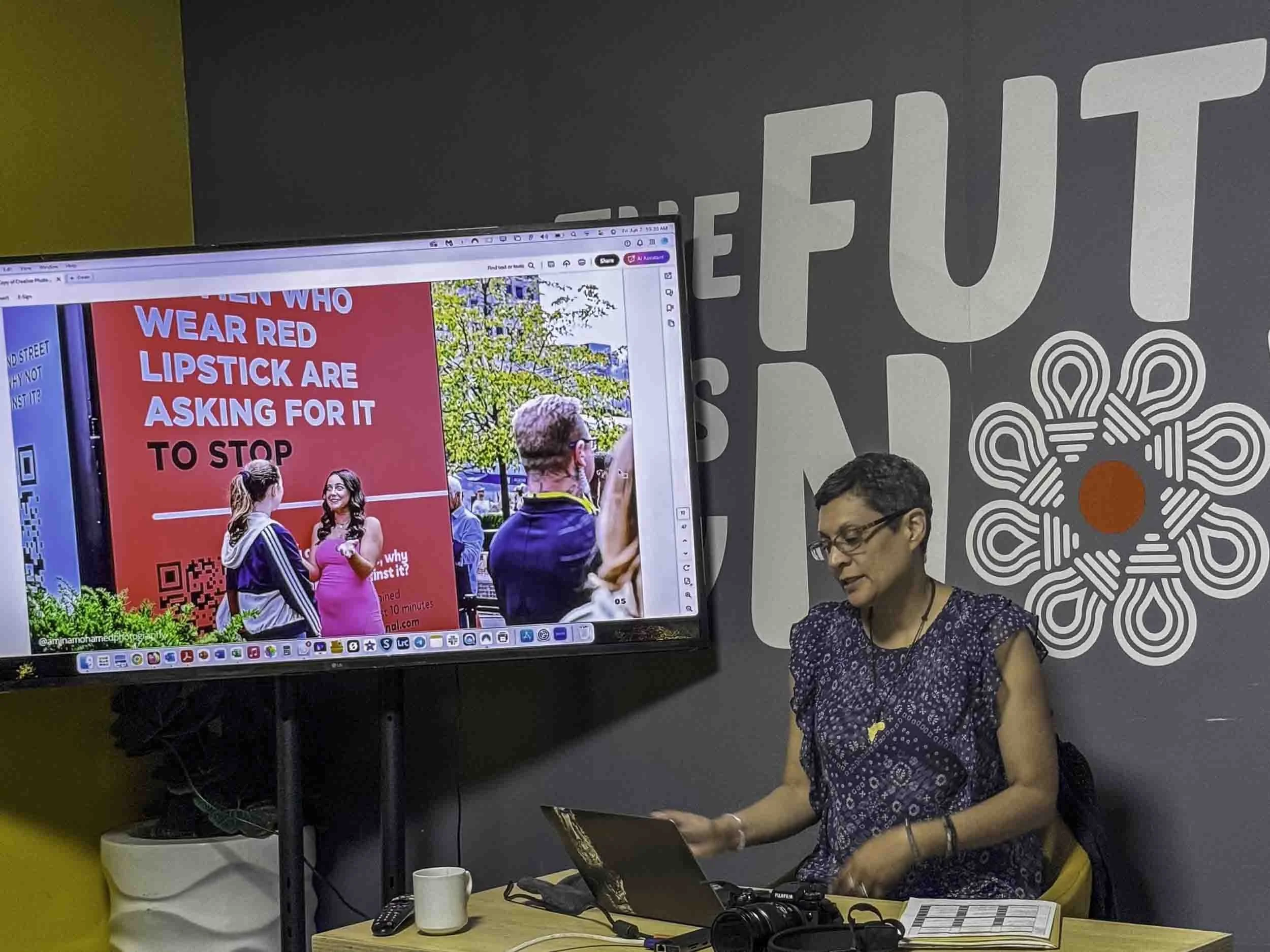 A woman with short dark hair and glasses wearing a dark patterned shirt, standing at a table with a laptop, a white mug, a camera, and other items, looking down and smiling. Behind her is a large screen displaying a colorful presentation slide with p