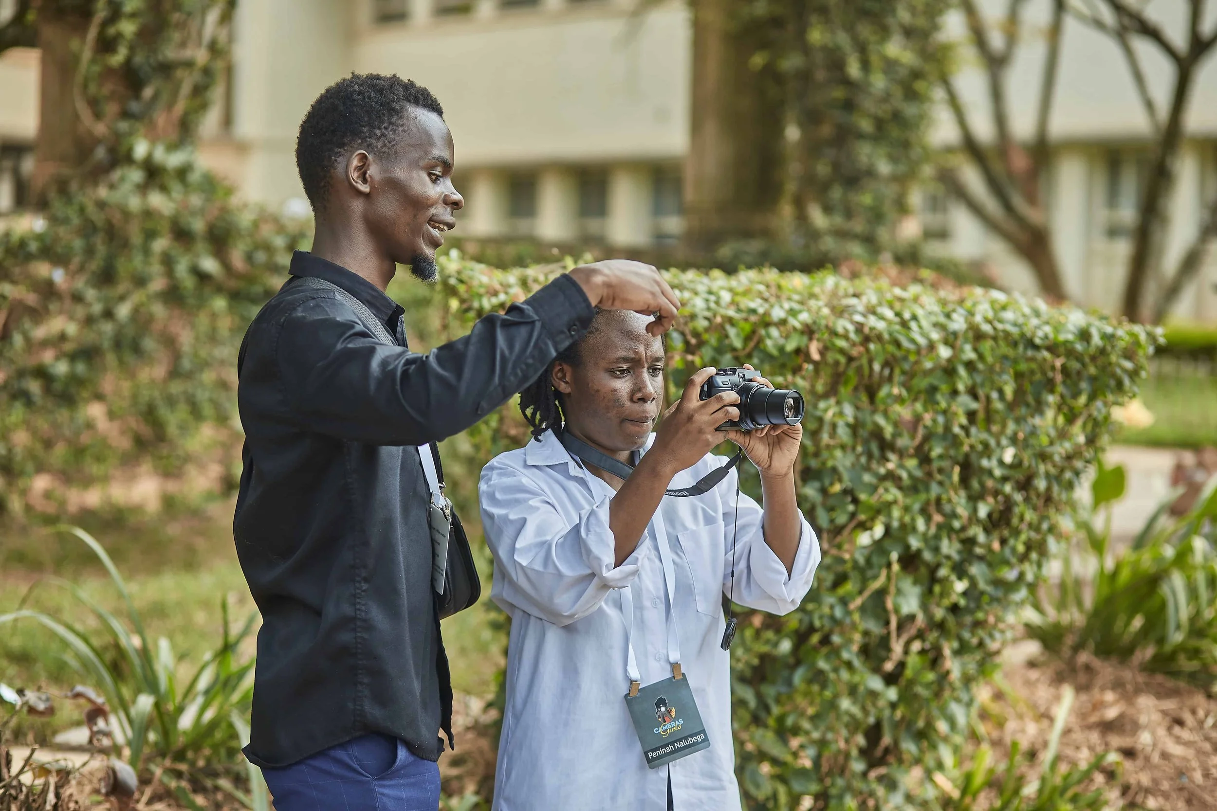 A man assists a woman with her camera outdoors, with greenery and trees in the background.