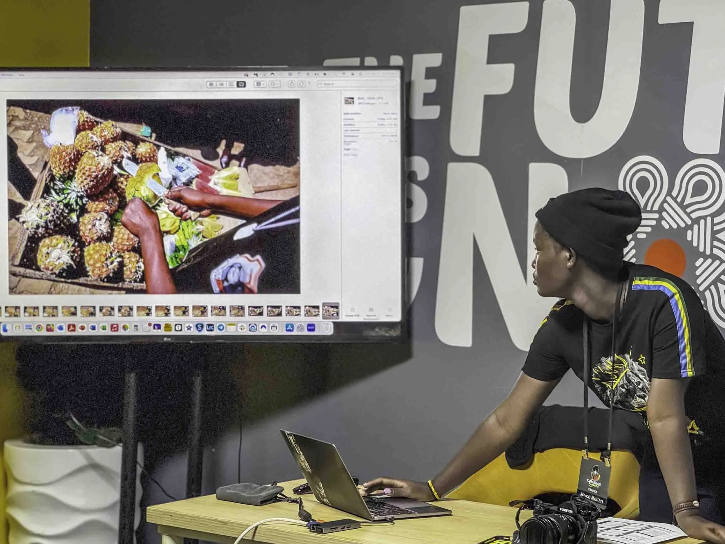 A person with a dark cap and a colorful t-shirt sitting at a desk with a laptop, a camera, and other items, looking at a large computer screen showing a photo of tropical fruits.