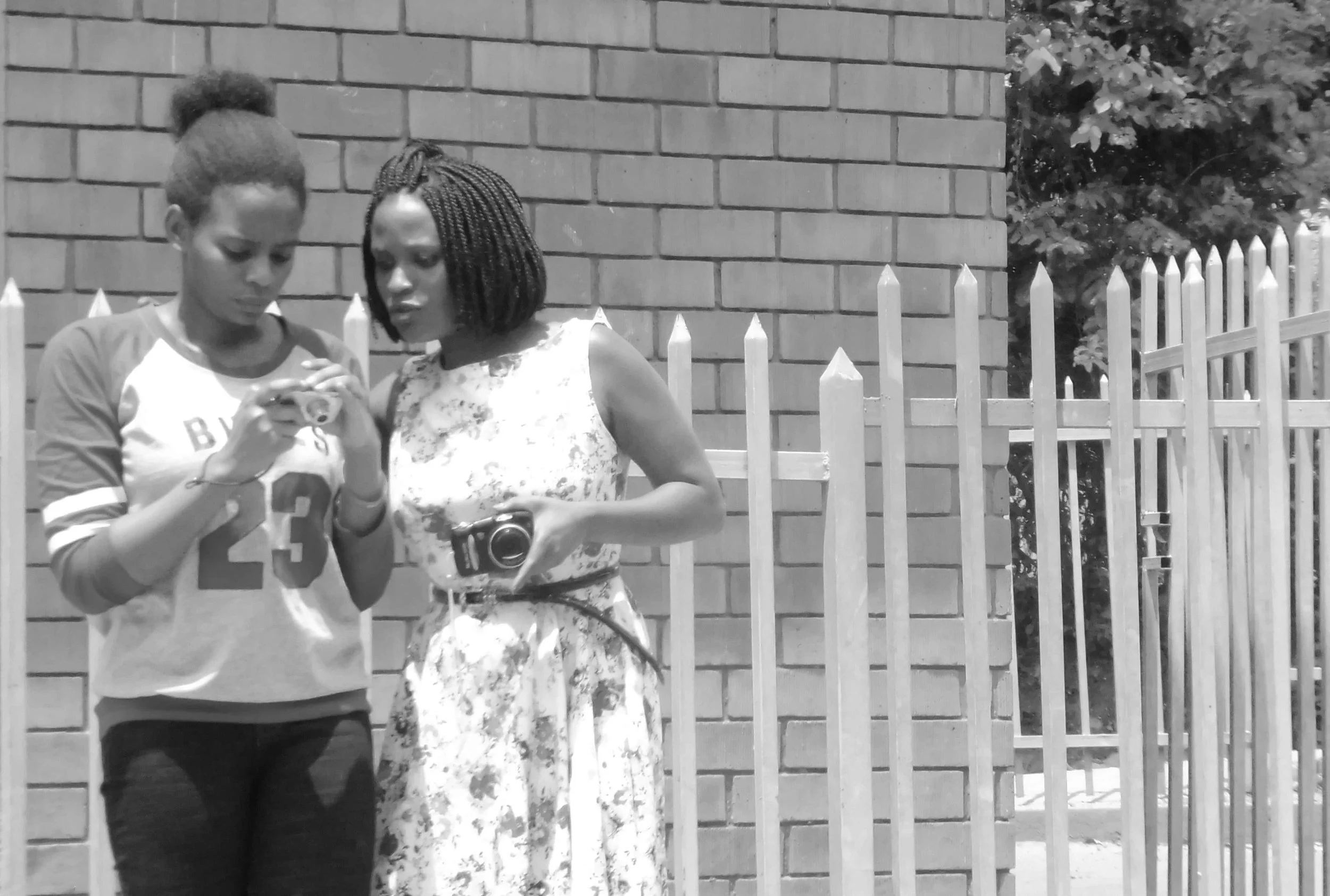 Two young women standing outdoors near a brick wall and white picket fence, looking at a camera or phone together.