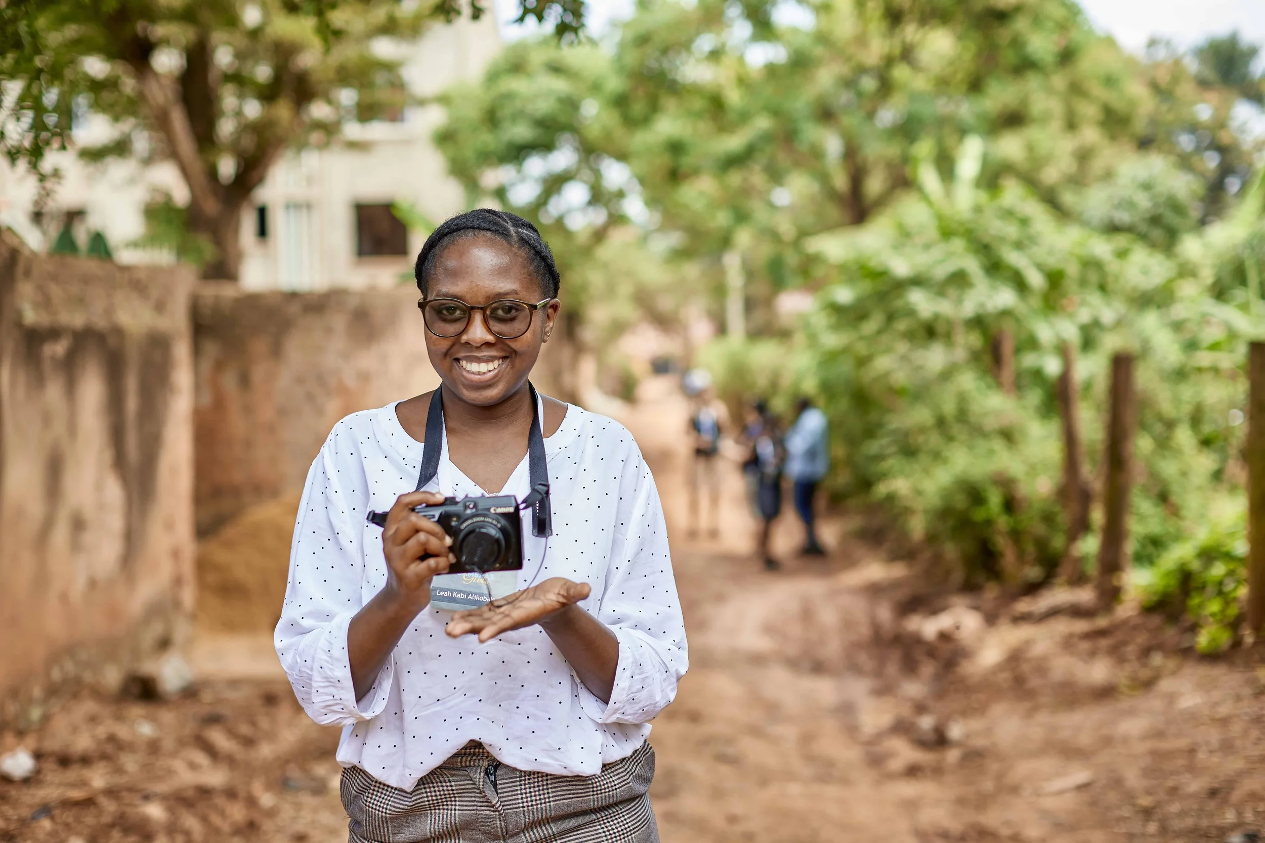 A young woman smiling and holding a camera outdoors with trees and a dirt path in the background.
