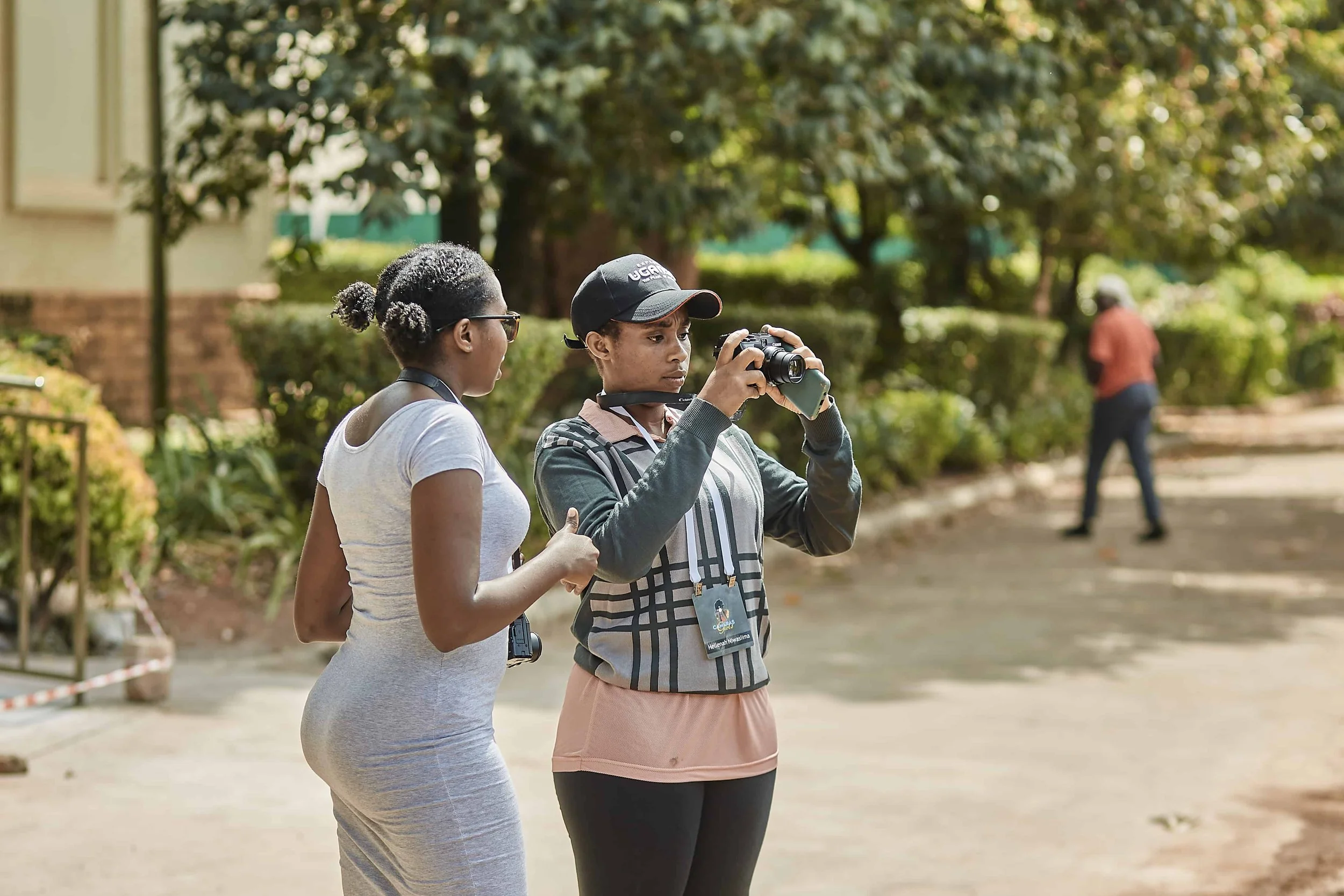 Two women standing outdoors, with trees and a person walking in the background. One woman is taking a photo with a camera, and the other woman is observing.