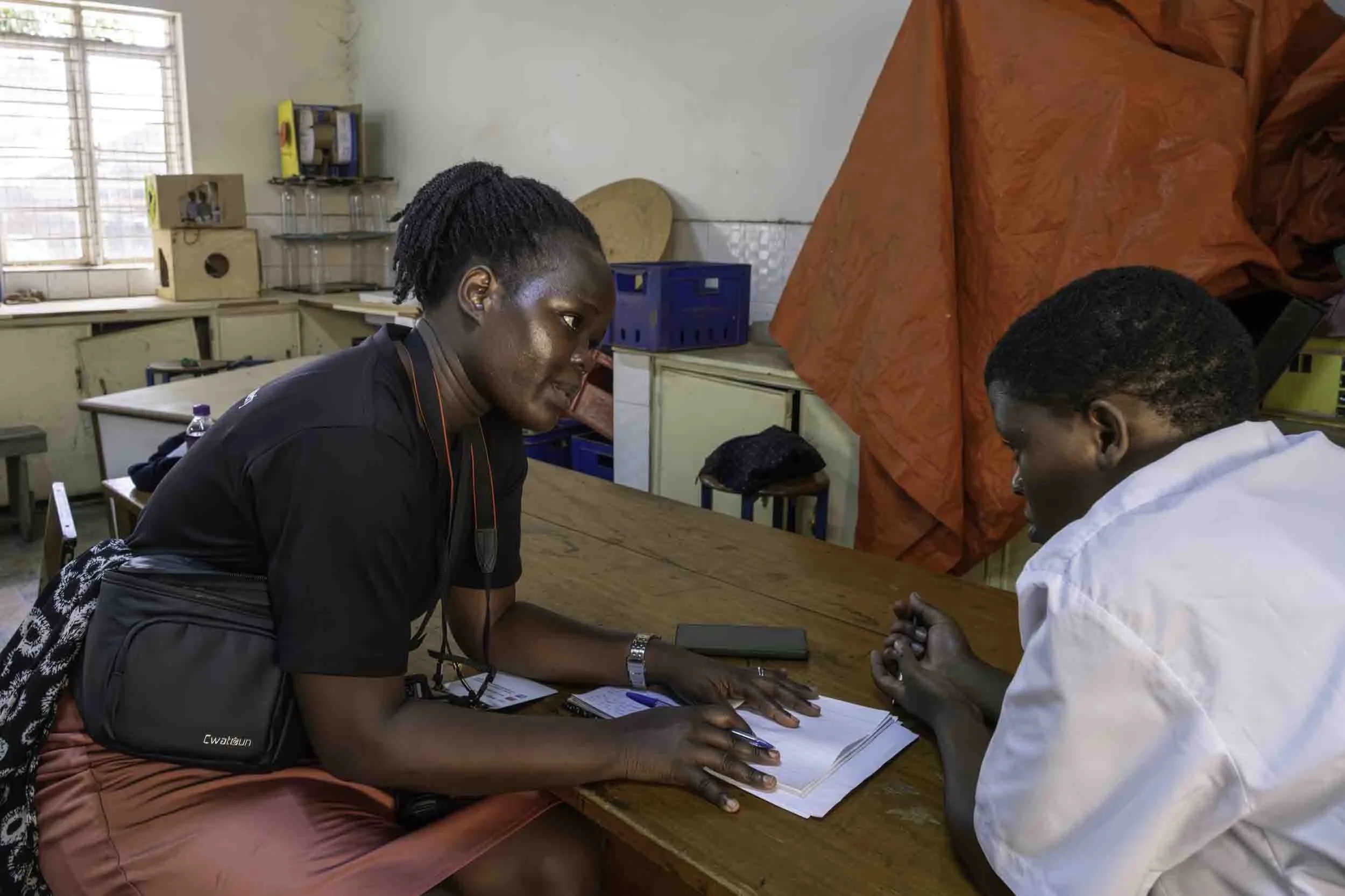 A healthcare worker is drawing blood from a patient's finger in a classroom or healthcare setting. The healthcare worker is wearing a black shirt and orange shorts, while the patient is wearing a white shirt. There is a woman with short, styled hair 