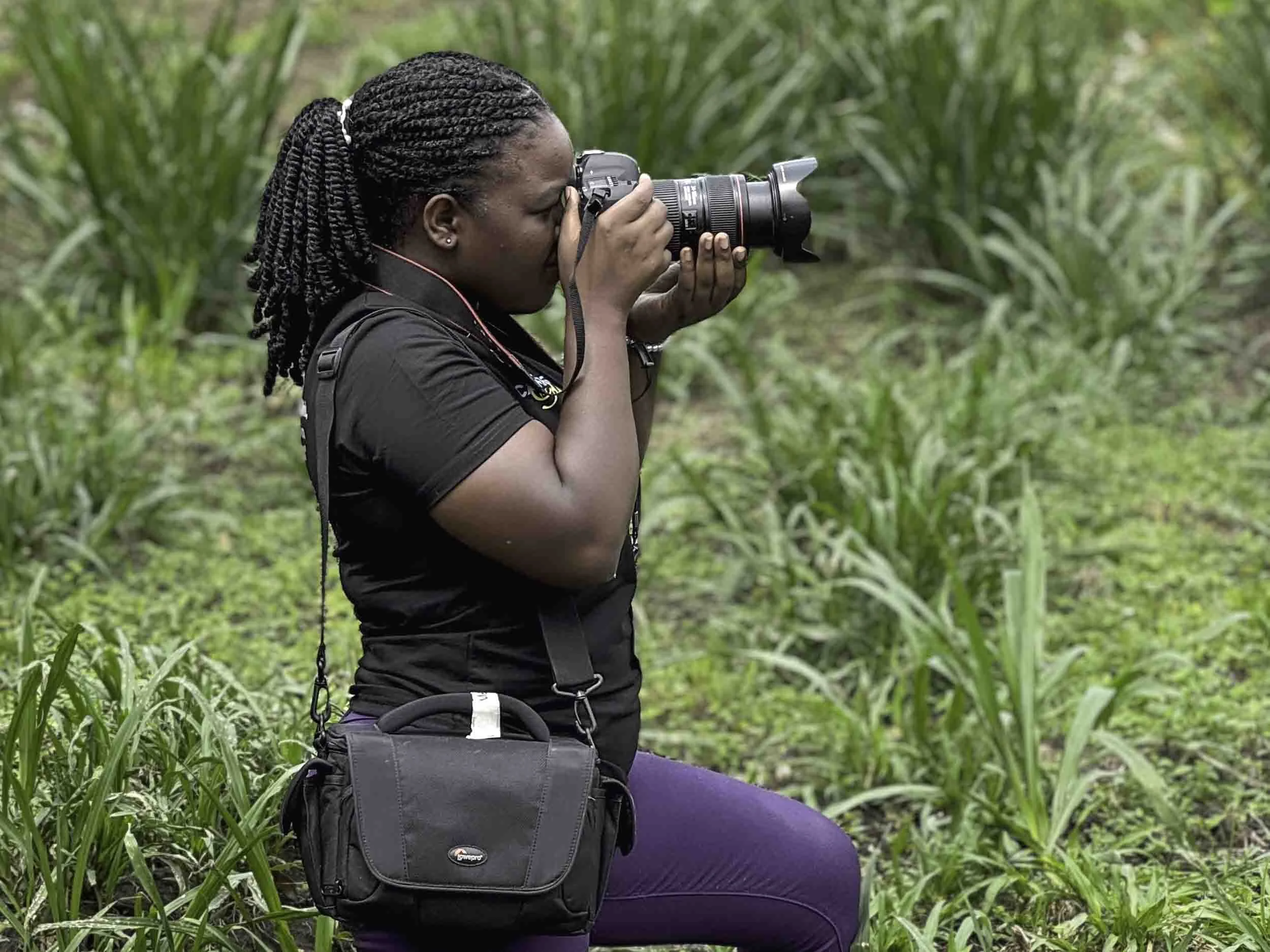 A woman with braided hair sitting outdoors among green tall grass or crops, taking a photograph with a DSLR camera. She is wearing a black shirt, purple pants, and has a black camera bag.